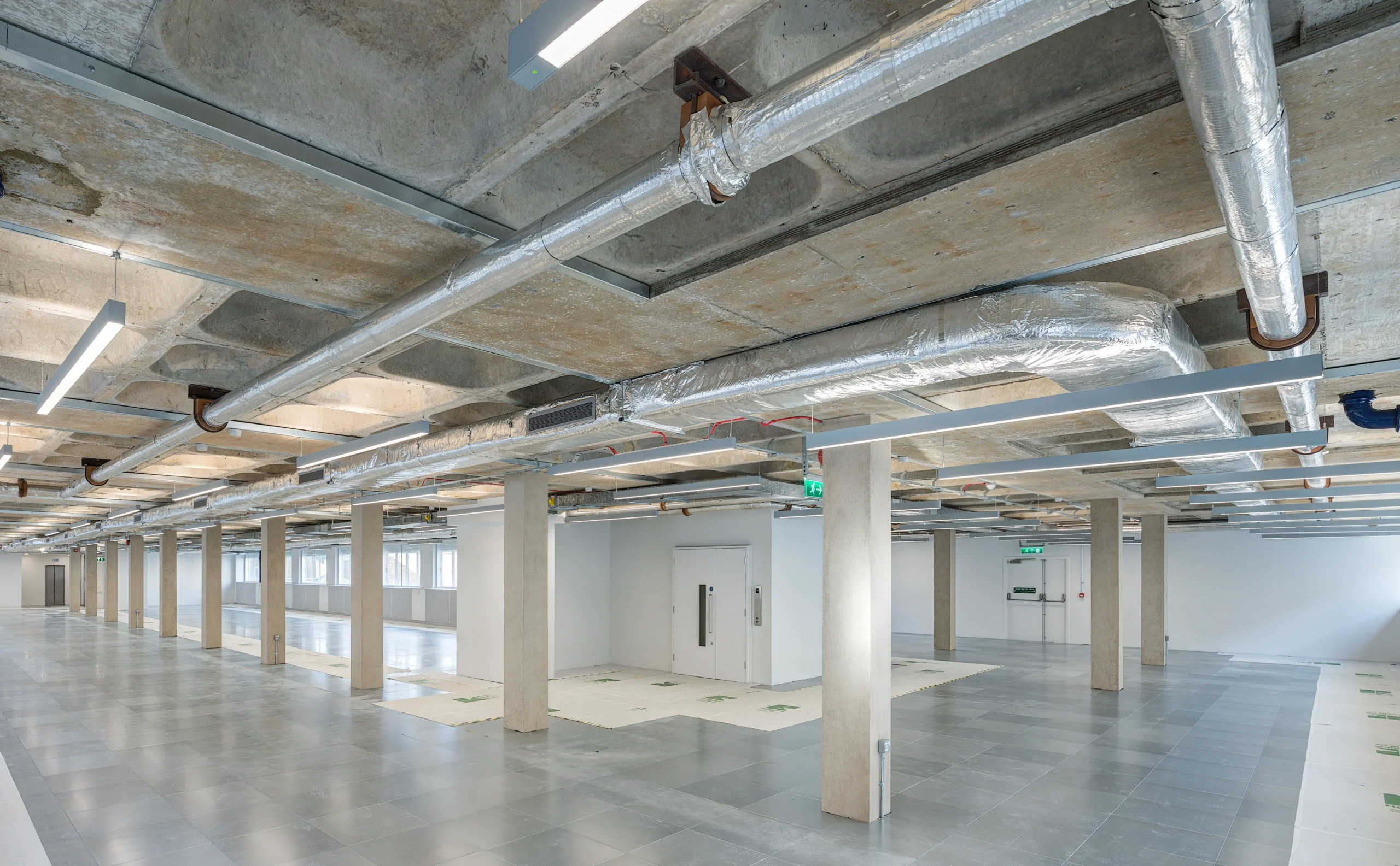 Interior view of Glen House showcasing an open-plan layout with exposed ceilings, concrete pillars, metal ductwork, and large windows. A staircase with glass doors is visible, connecting the workspace to other areas.