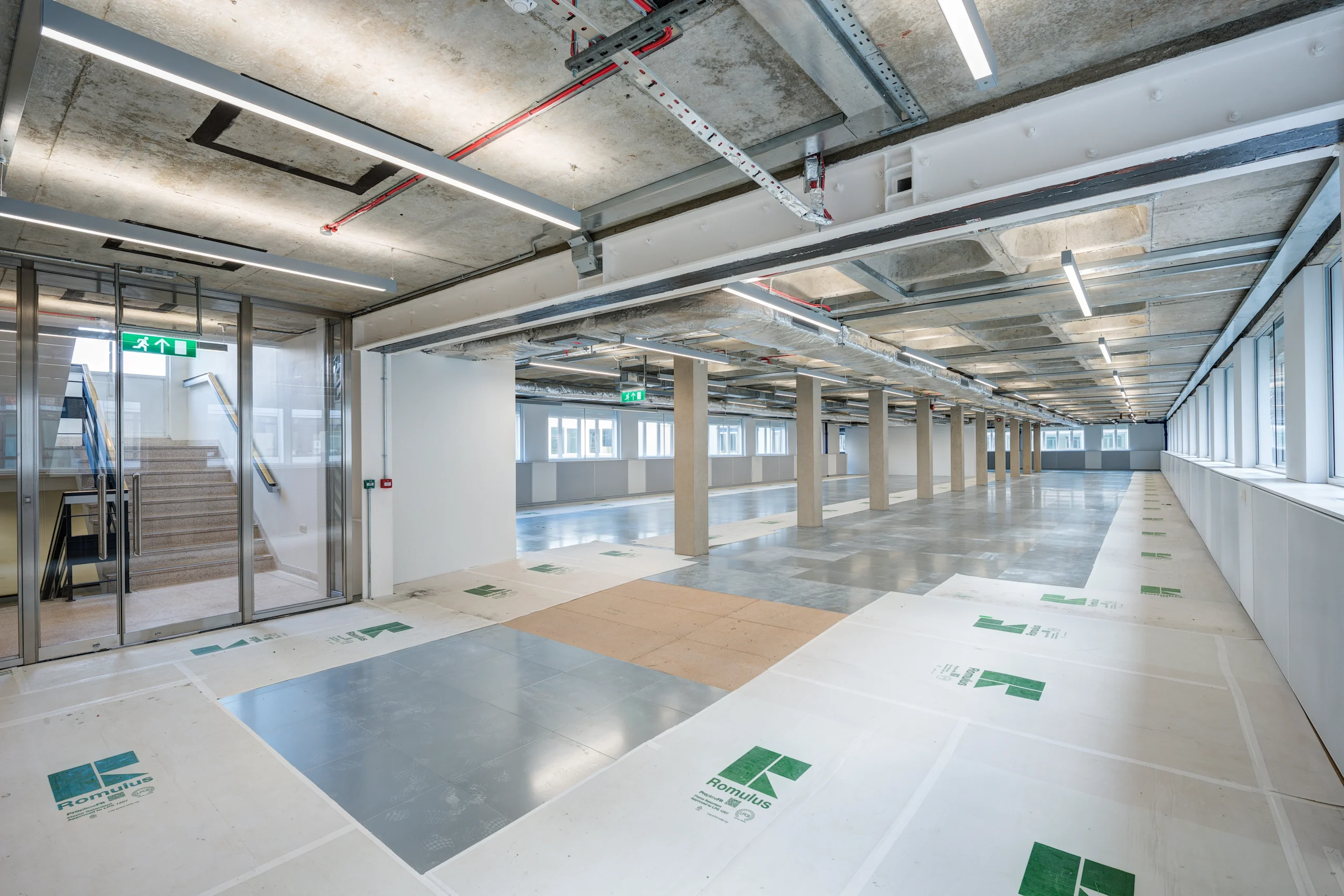 Interior view of Glen House showcasing an open-plan layout with exposed ceilings, concrete pillars, metal ductwork, and large windows. A staircase with glass doors is visible, connecting the workspace to other areas.