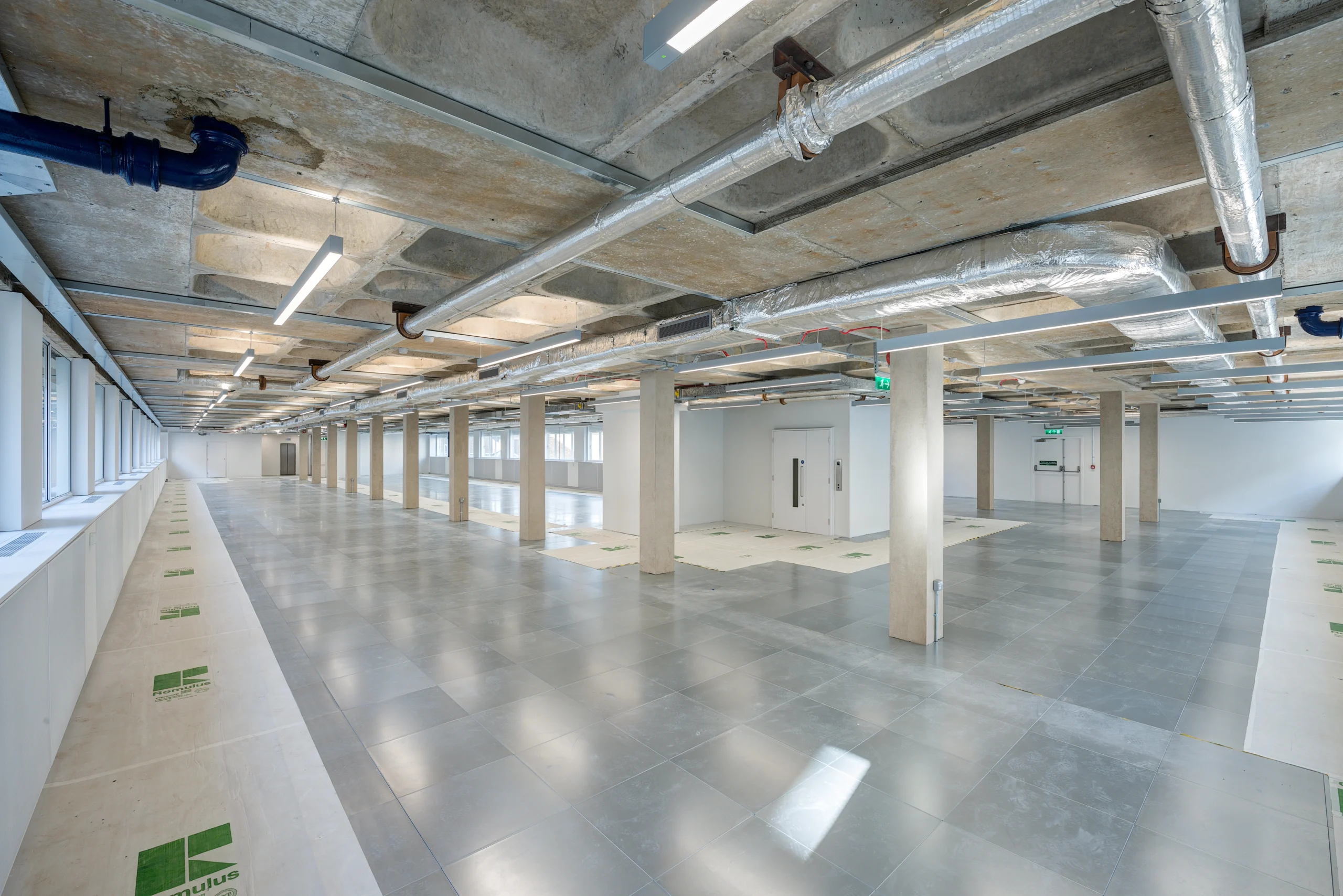 Open-plan industrial space at Glen House with exposed ceilings, concrete pillars, metal ductwork, and large windows providing natural light.