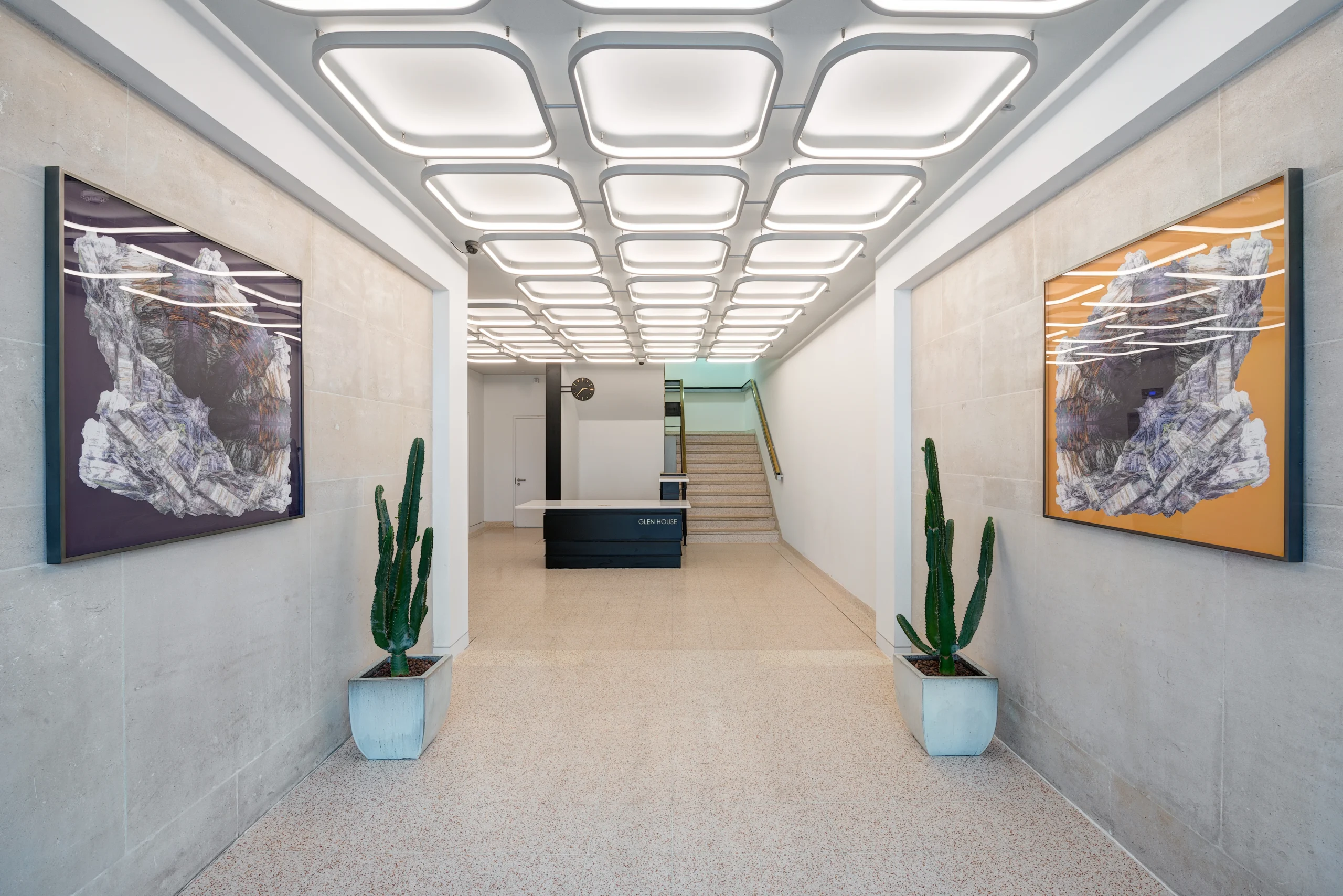 Modern lobby at Glen House with illuminated ceiling panels, abstract artwork on the walls, and potted cacti on either side, leading to a sleek reception desk and stairs.