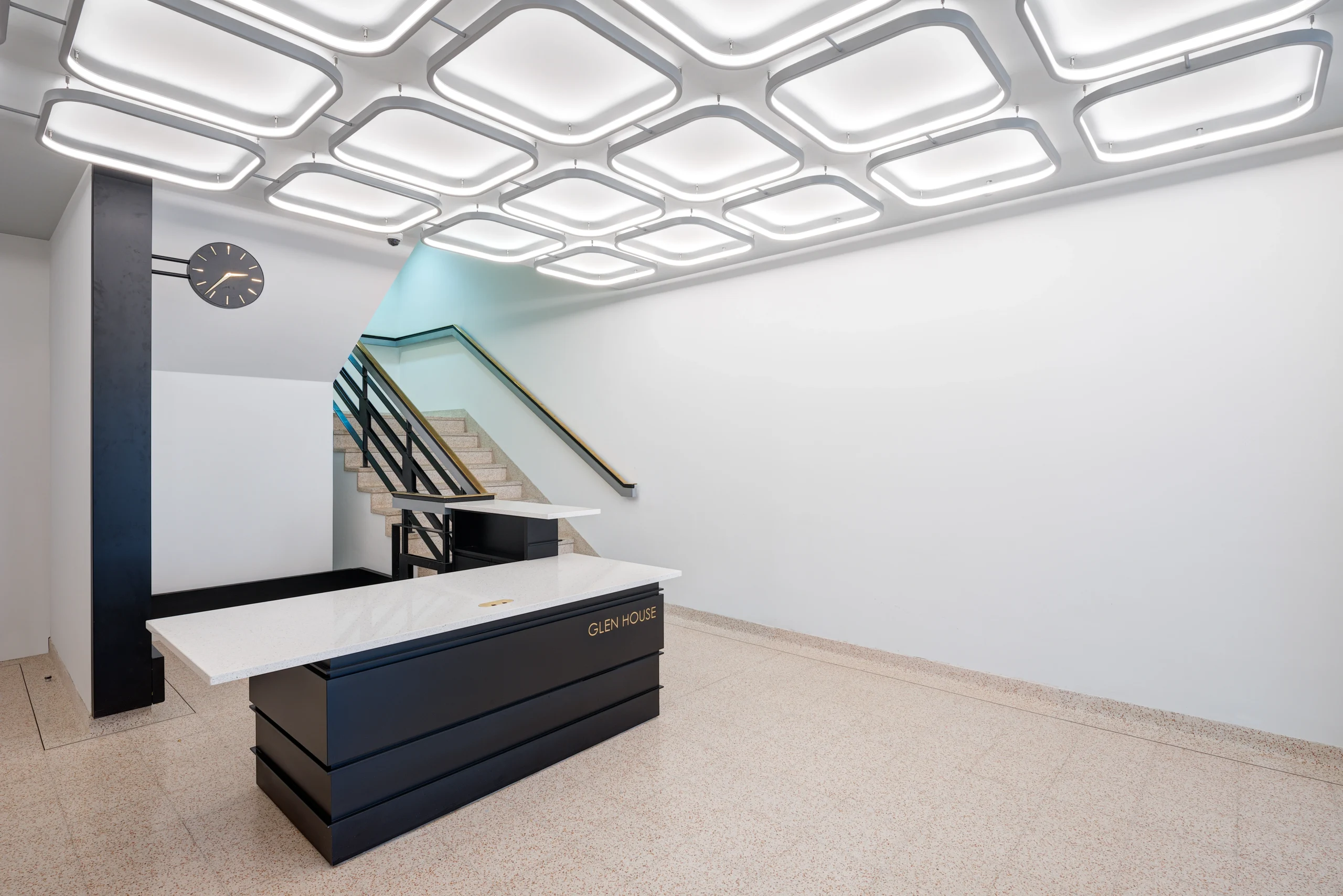 Reception area at Glen House featuring a modern black desk with 'Glen House' lettering, illuminated ceiling panels, and a staircase with sleek metal railings leading to the upper floor.