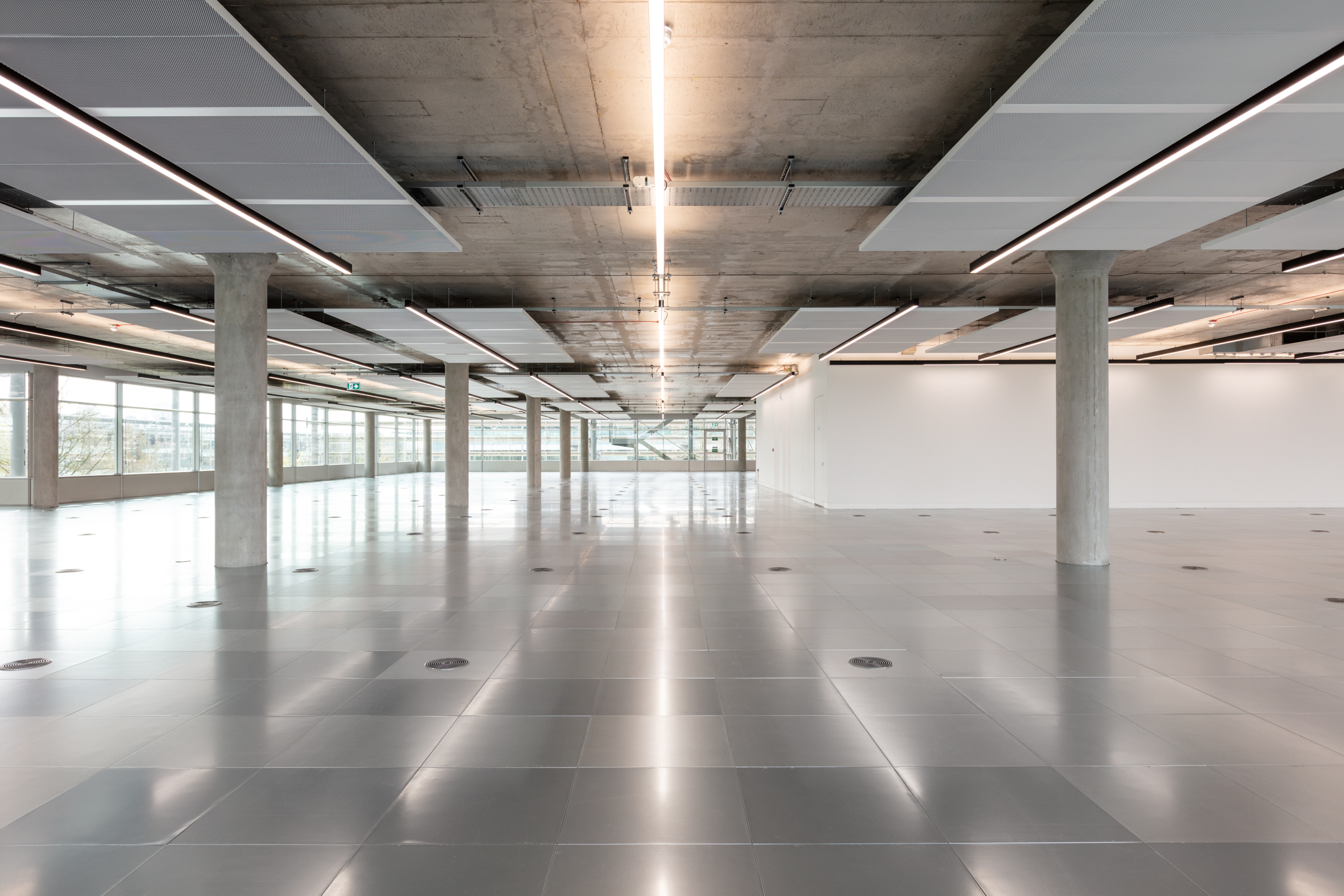 Spacious Interior of Chiswick Business Park Building 5: A spacious and modern open-plan interior of Chiswick Building 5, featuring concrete columns and ceiling with exposed panels and integrated lighting. The floor is polished, reflecting the lights above.