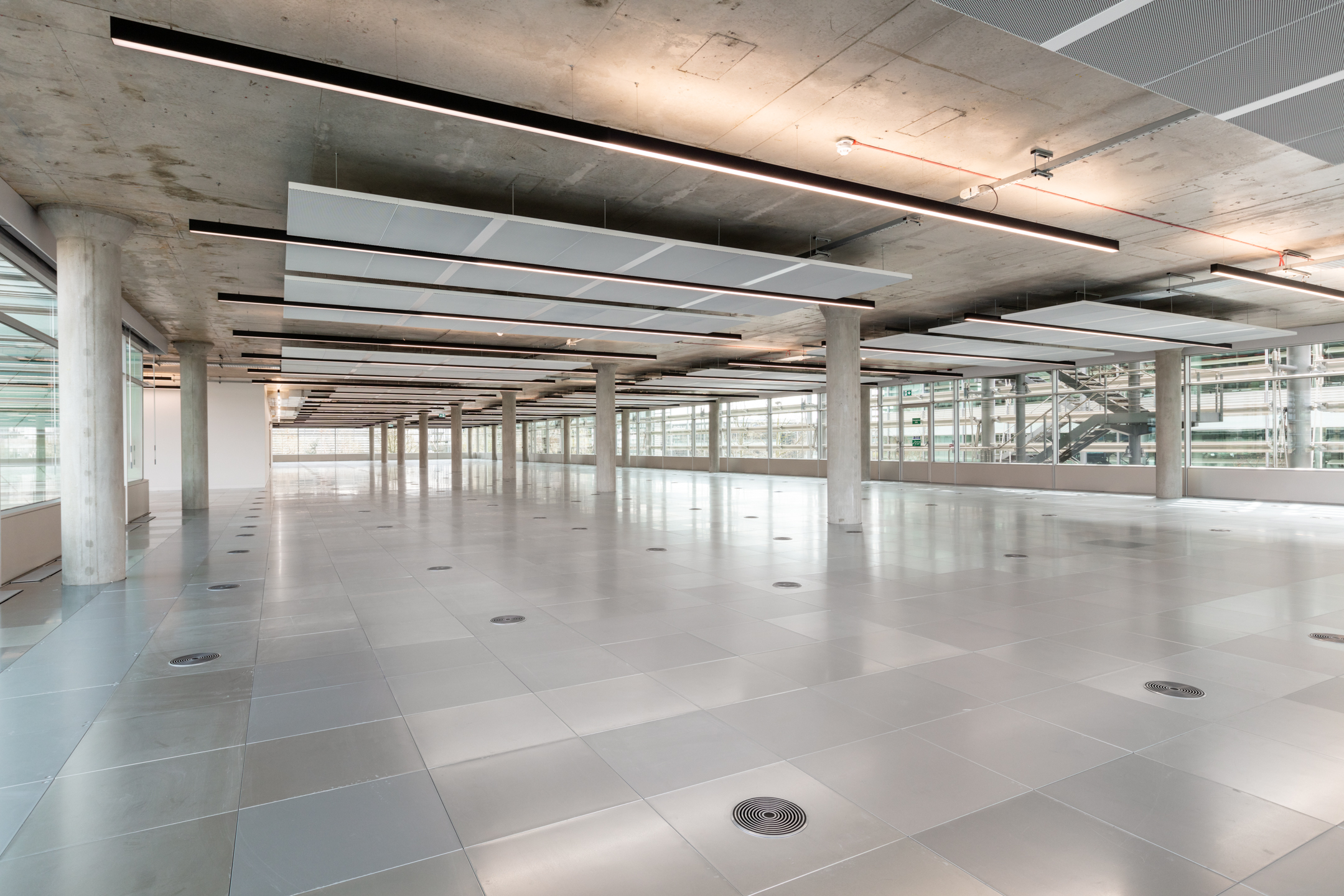 Modern Open Space Design in Chiswick Building 5: The interior of Chiswick Building 5, showcasing a large open space with reflective flooring, concrete columns, and ceiling panels. The glass walls offer a panoramic view of the surrounding area.