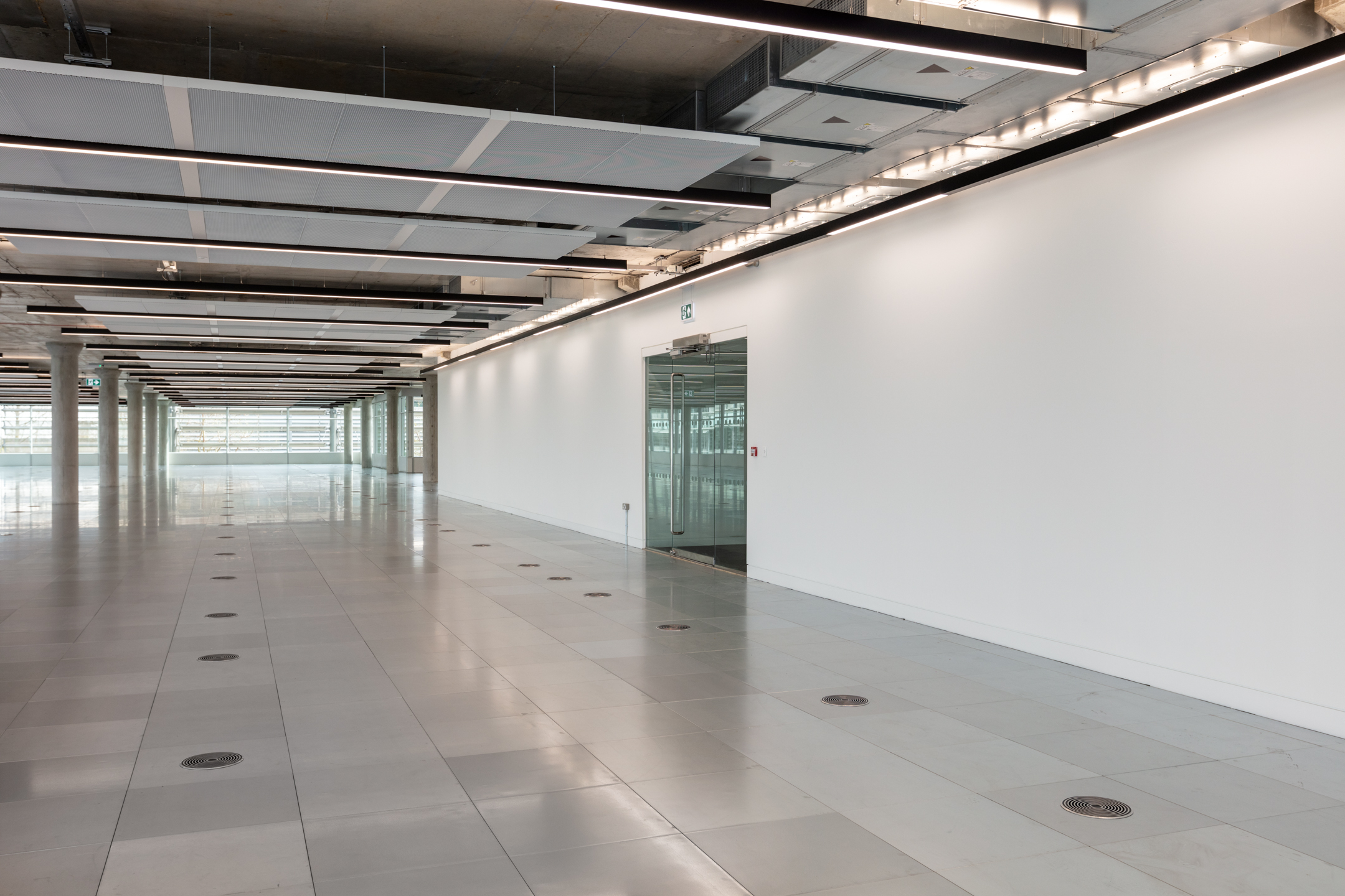 Hallway Leading to Glass-Panelled Exit in Chiswick Building 5: A hallway inside Chiswick Business Park Building 5, leading towards a glass-panelled exit. The walls are white, with integrated ceiling lights running along the length of the corridor.