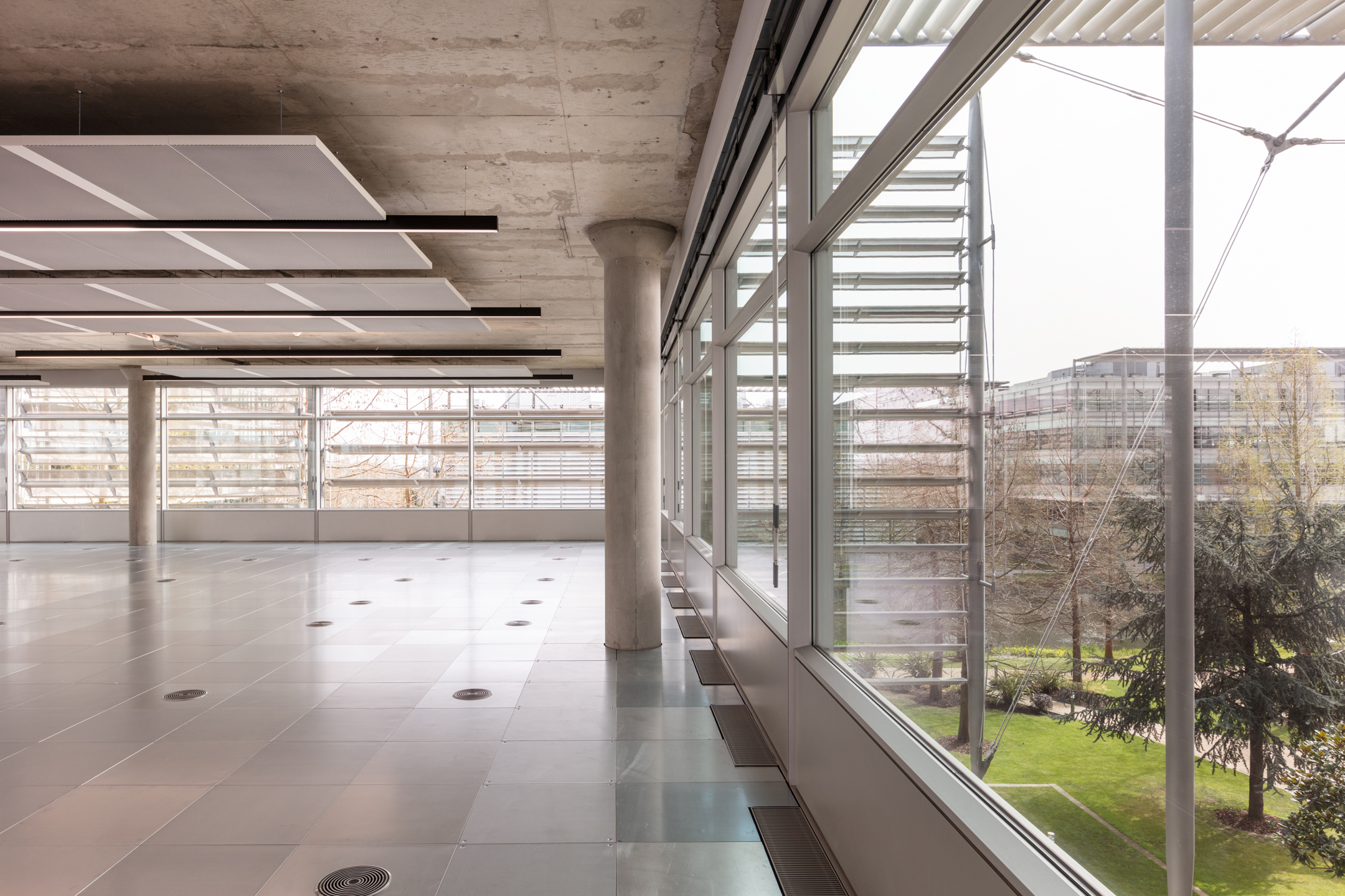 Bright Workspace Overlooking the Exterior of Chiswick Building 5: A bright workspace inside Chiswick Business Park Building 5, with large windows and views of the exterior landscape, including trees and modern architecture outside. The flooring is metallic, and the ceiling is fitted with panels and lighting fixtures.