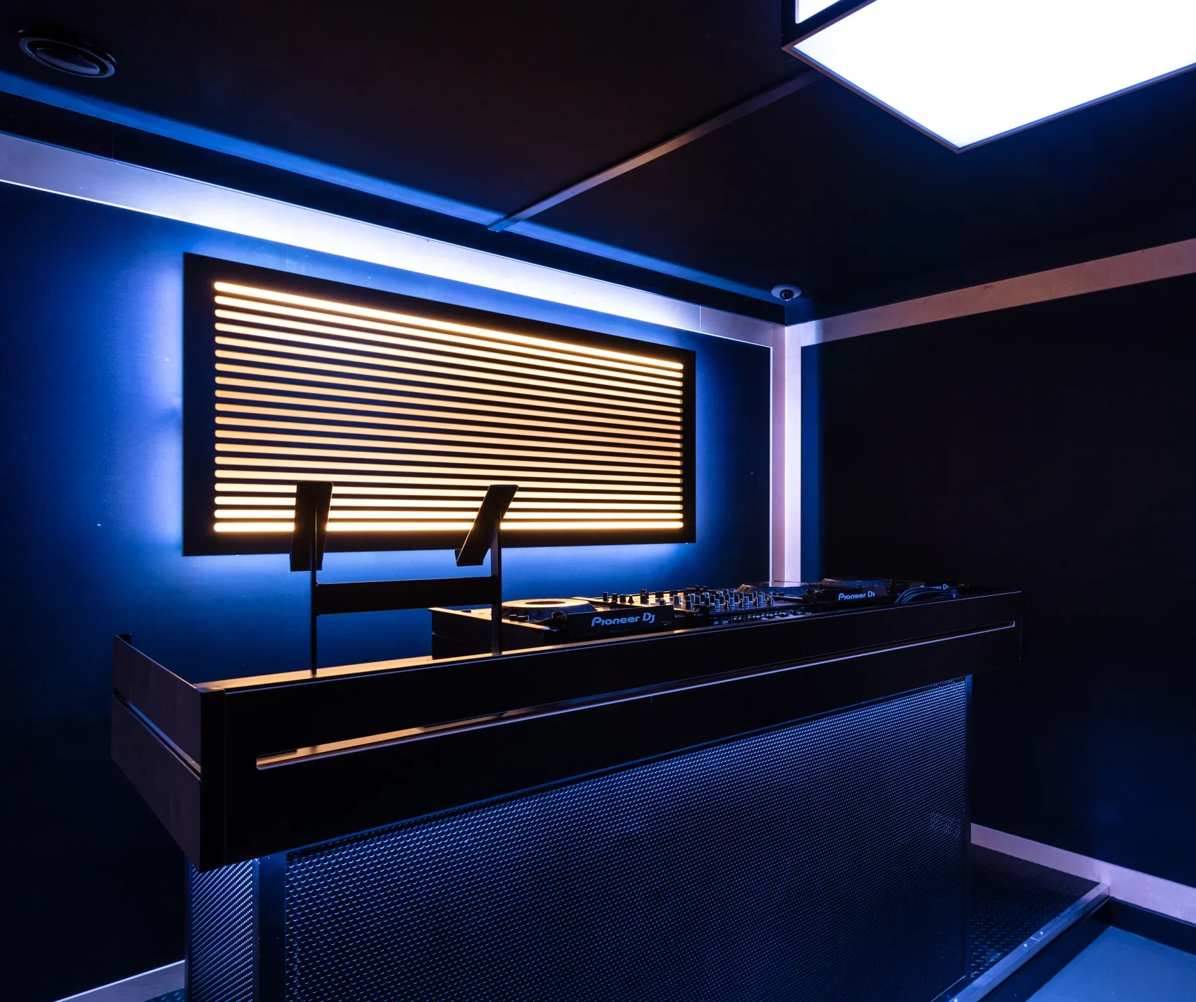 A DJ booth at Pirate Studio, featuring a sleek black console with a pair of Pioneer DJ decks. The backdrop is illuminated with horizontal light strips in a dark, ambient room.