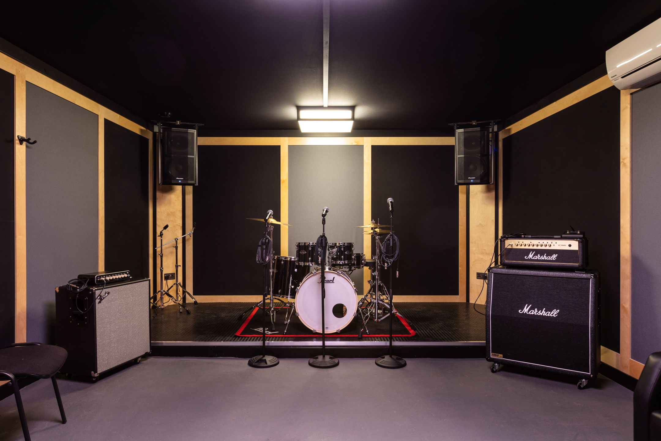 A rehearsal room at Pirate Studio, equipped with a drum kit on a small stage, two microphones on stands, a Marshall amplifier, and wall-mounted speakers in a soundproofed environment.