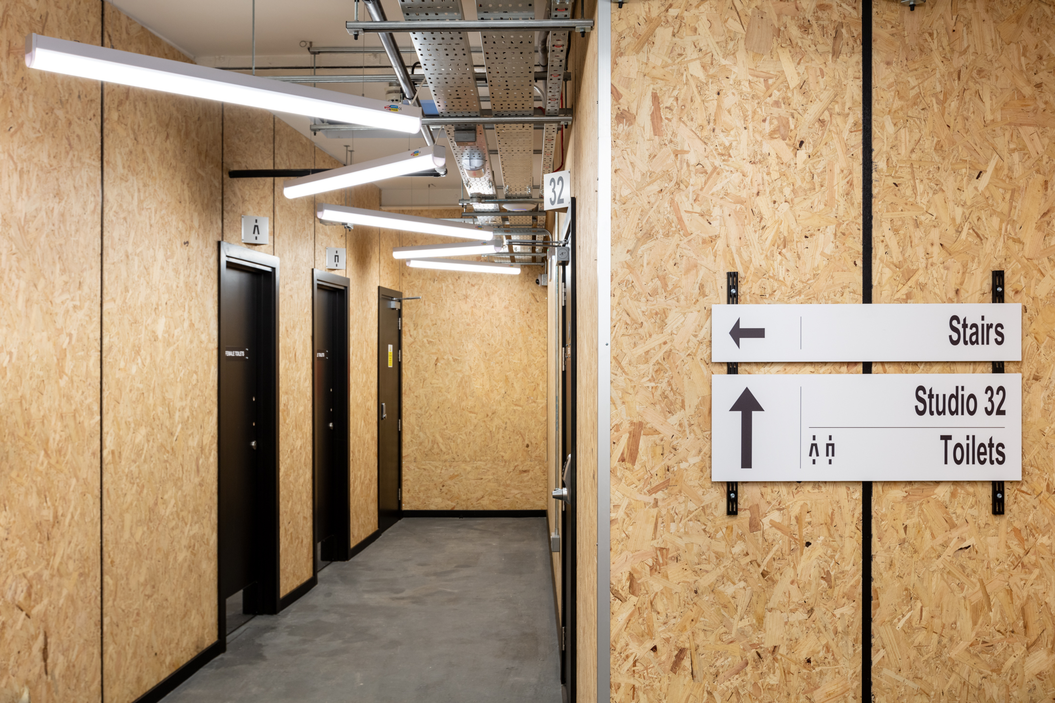 A corridor at Pirate Studio featuring wood-panelled walls, overhead exposed lighting, and directional signage pointing towards Studio 32, stairs, and toilets.