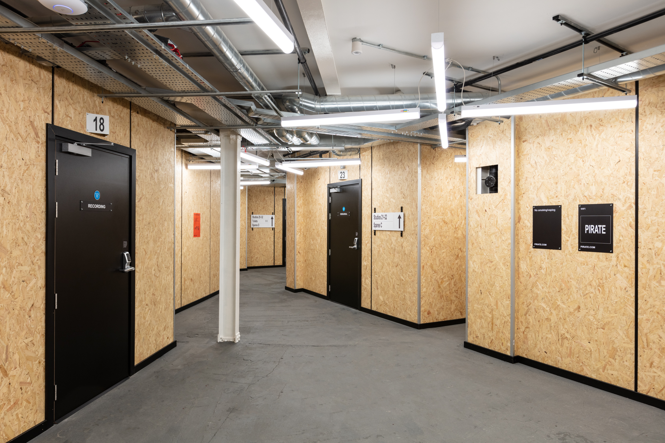 Interior corridor at Pirate Studios, featuring industrial-inspired design with exposed piping, OSB wall panelling, and directional signage for various recording and rehearsal rooms.