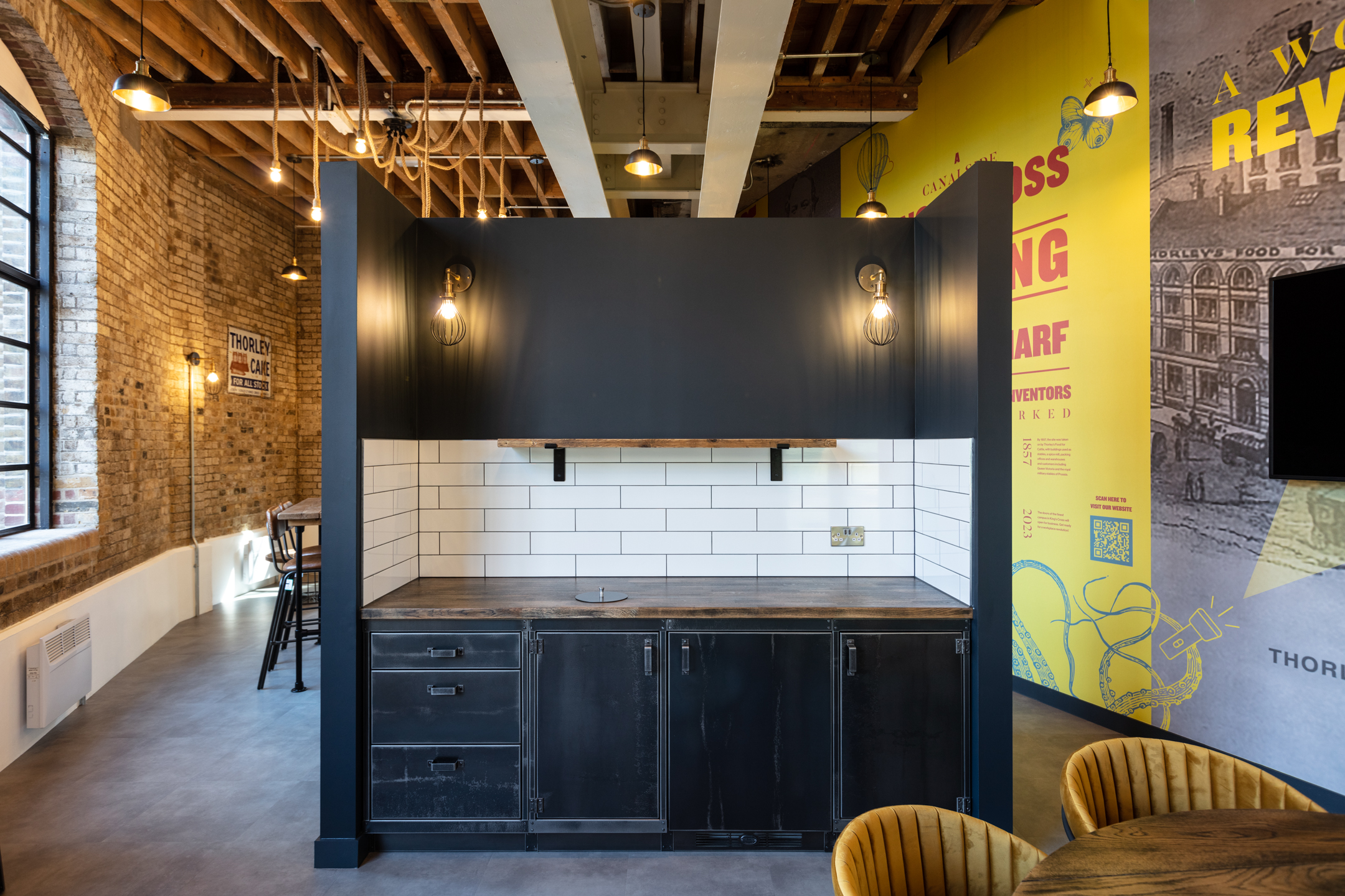 Industrial-Style Teapoint at Regent's Wharf: A small teapoint area at Regent's Wharf with black metal cabinets, a wooden countertop, and a white subway tile backsplash. The ceiling features rustic rope lighting.