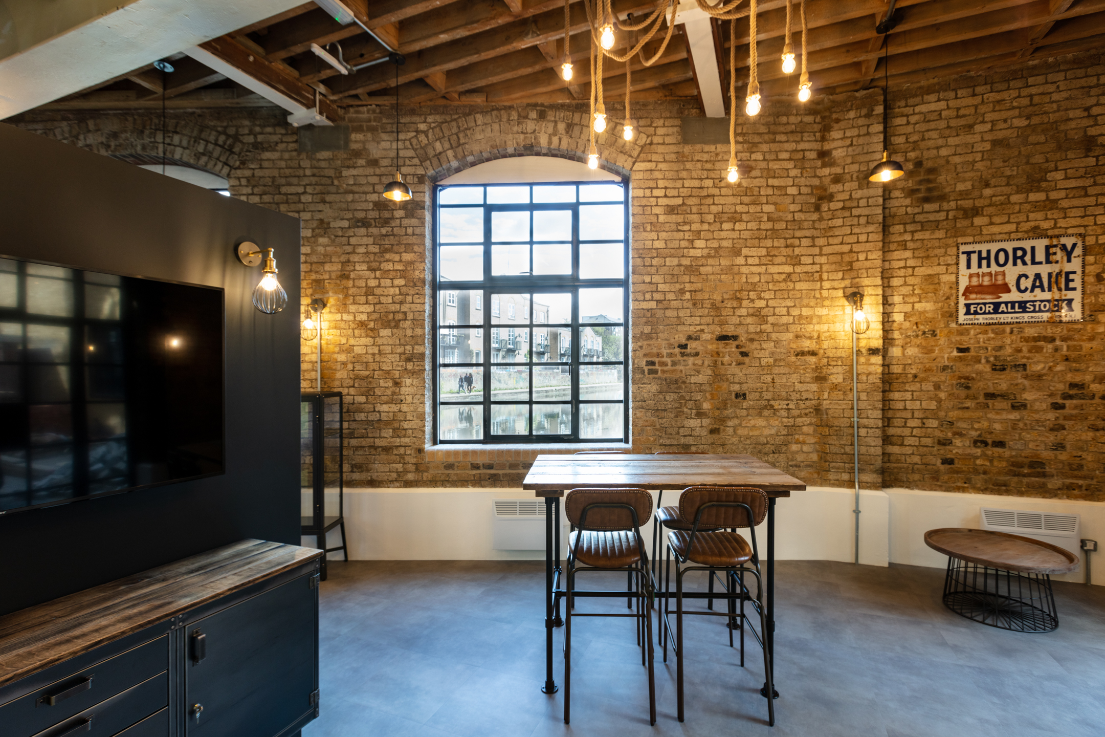 Brick Interior with Classic Windows at Regent's Wharf: Interior view of Regent's Wharf showing a classic brick wall with tall arched windows. There is a wooden table with stools placed in the middle, and vintage lighting hanging from the ceiling.