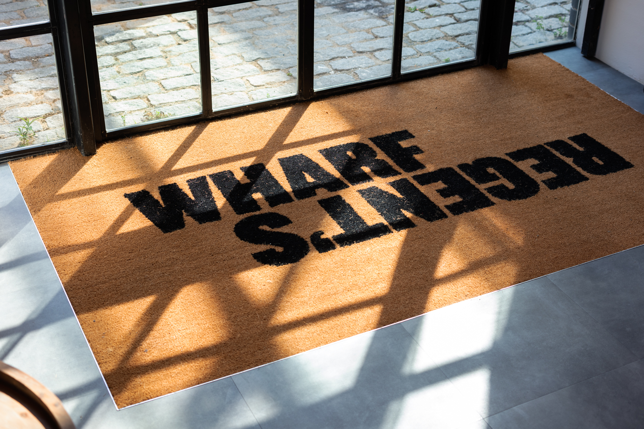 Regent's Wharf Branded Doormat by the Entrance: Regent's Wharf branded doormat by the entrance, with natural light streaming through the windows and casting shadows on the floor.