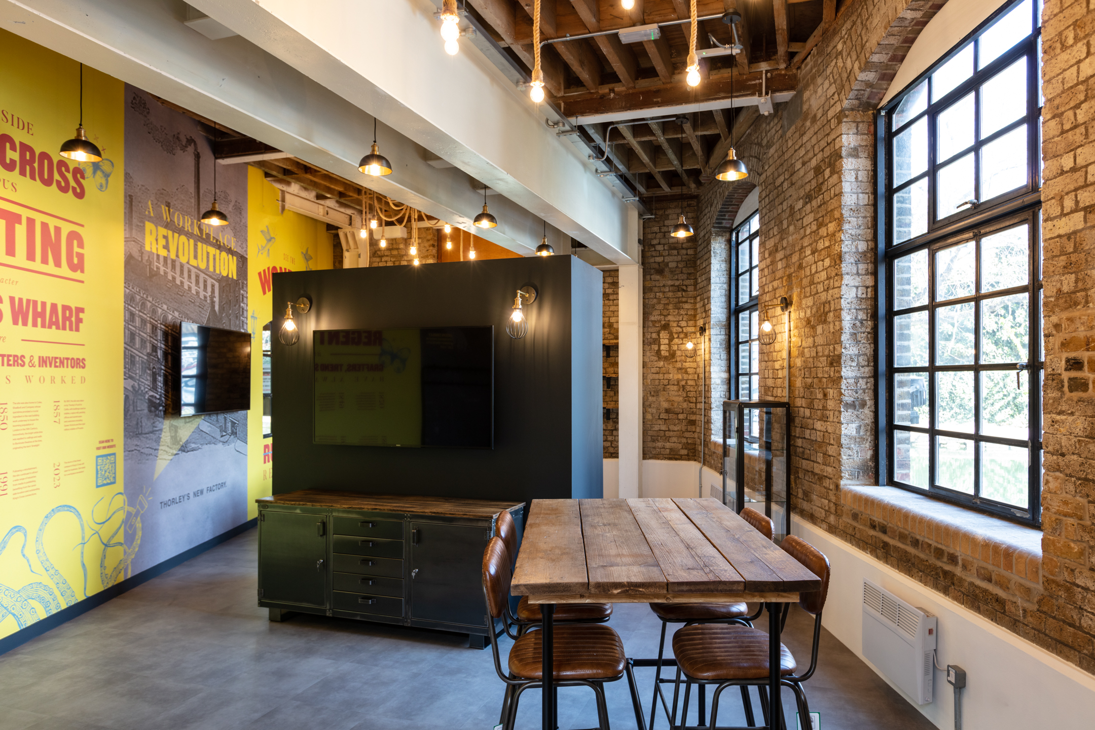 Open Space with Rustic Charm at Regent's Wharf: A meeting space at Regent's Wharf featuring industrial-style décor with exposed brick walls, metal finishes, and pendant lighting. A wooden table and vintage-style chairs provide seating, with bold wall graphics in the background.