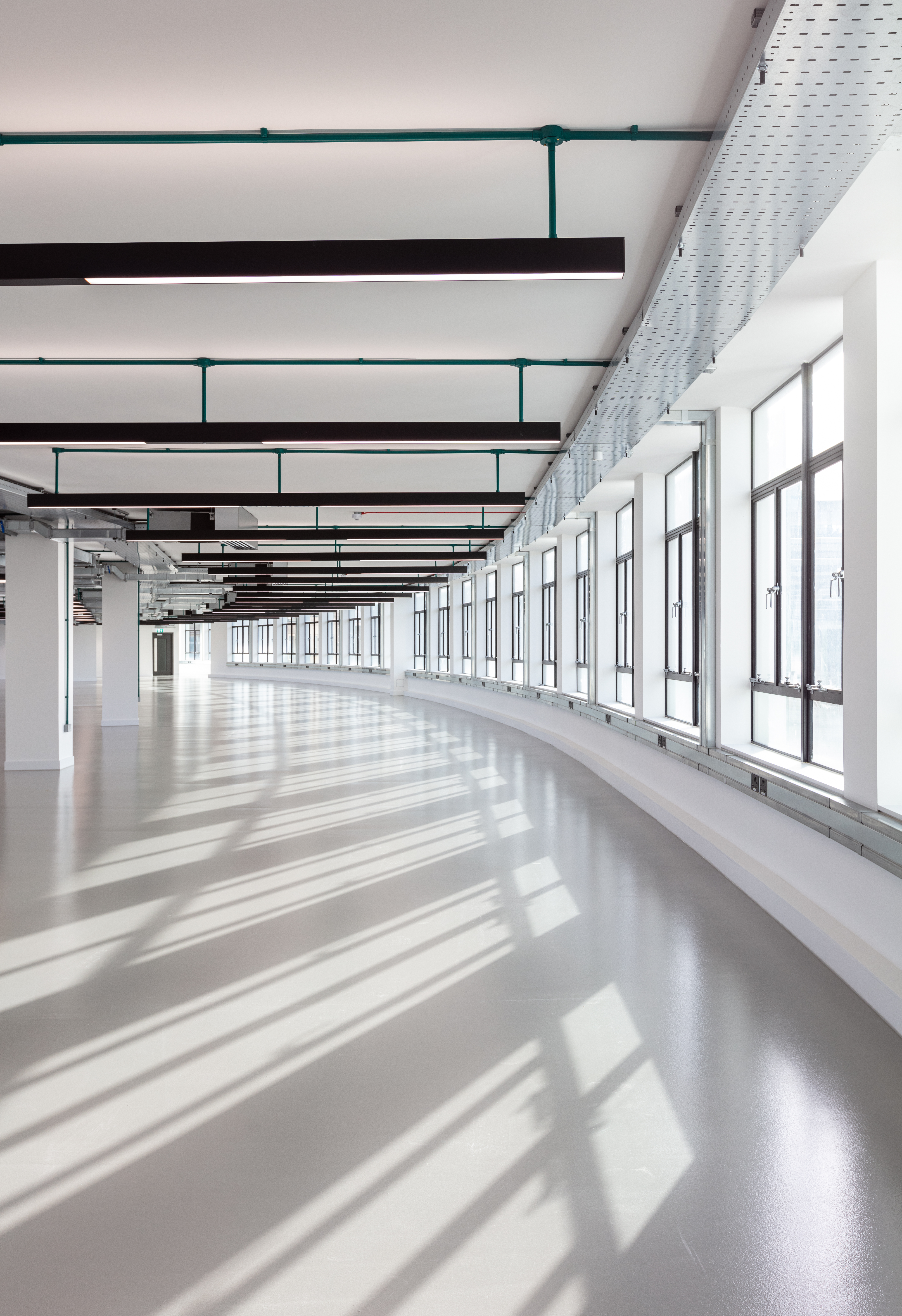 Bright, open-plan space within the Programme Building, featuring a curved wall of large windows casting geometric shadows across the polished floor, with exposed ceiling pipes and modern lighting.