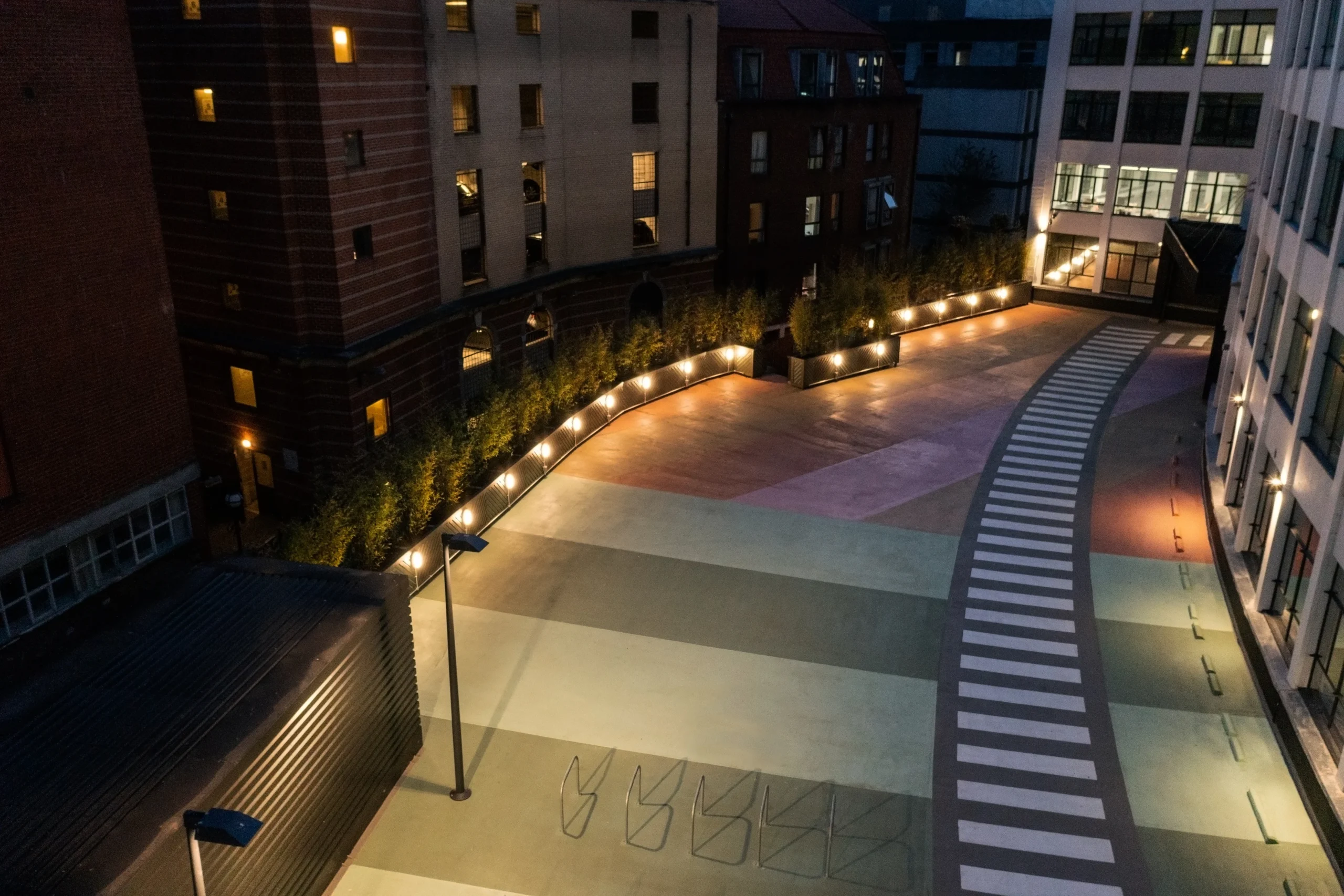 Evening view of the courtyard at the Programme Building, featuring well-lit pathways, plant-lined borders, and a striped walkway pattern that adds a dynamic visual element to the open space.