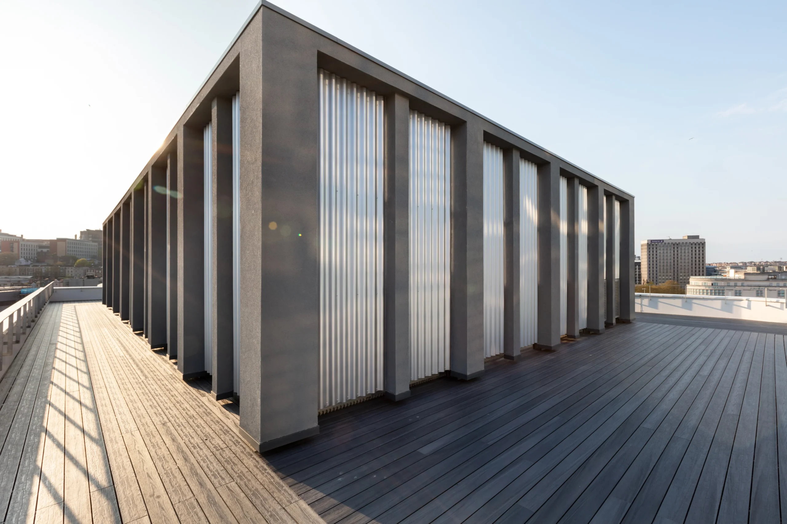 Sunlit rooftop deck of the Programme Building, showcasing a modern structure with evenly spaced columns and corrugated panels, providing a panoramic view of the surrounding cityscape.