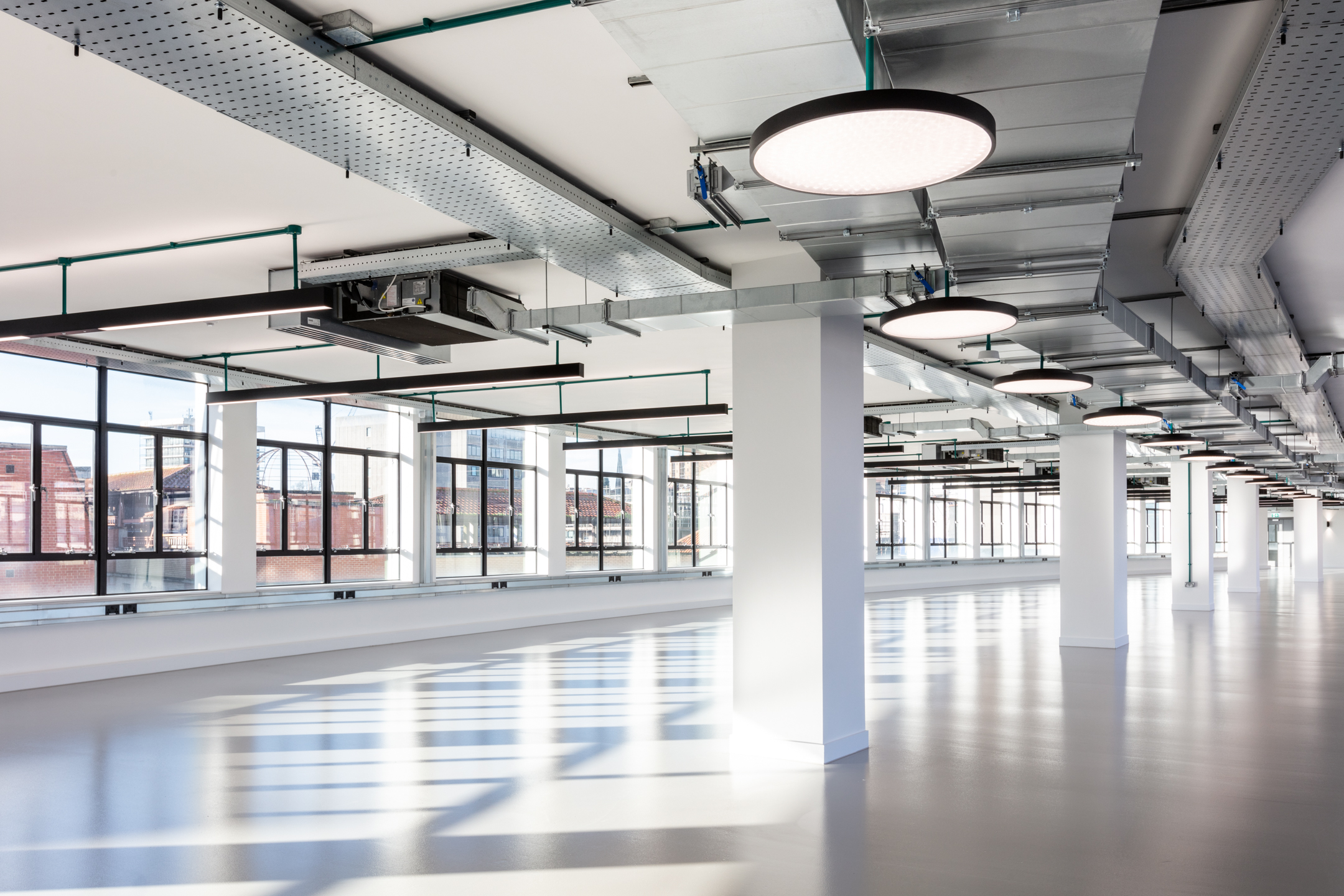 A spacious open-plan office space at Programme Building with large windows allowing natural light to flood the room. Exposed ductwork and industrial-style lighting fixtures add to the contemporary design.