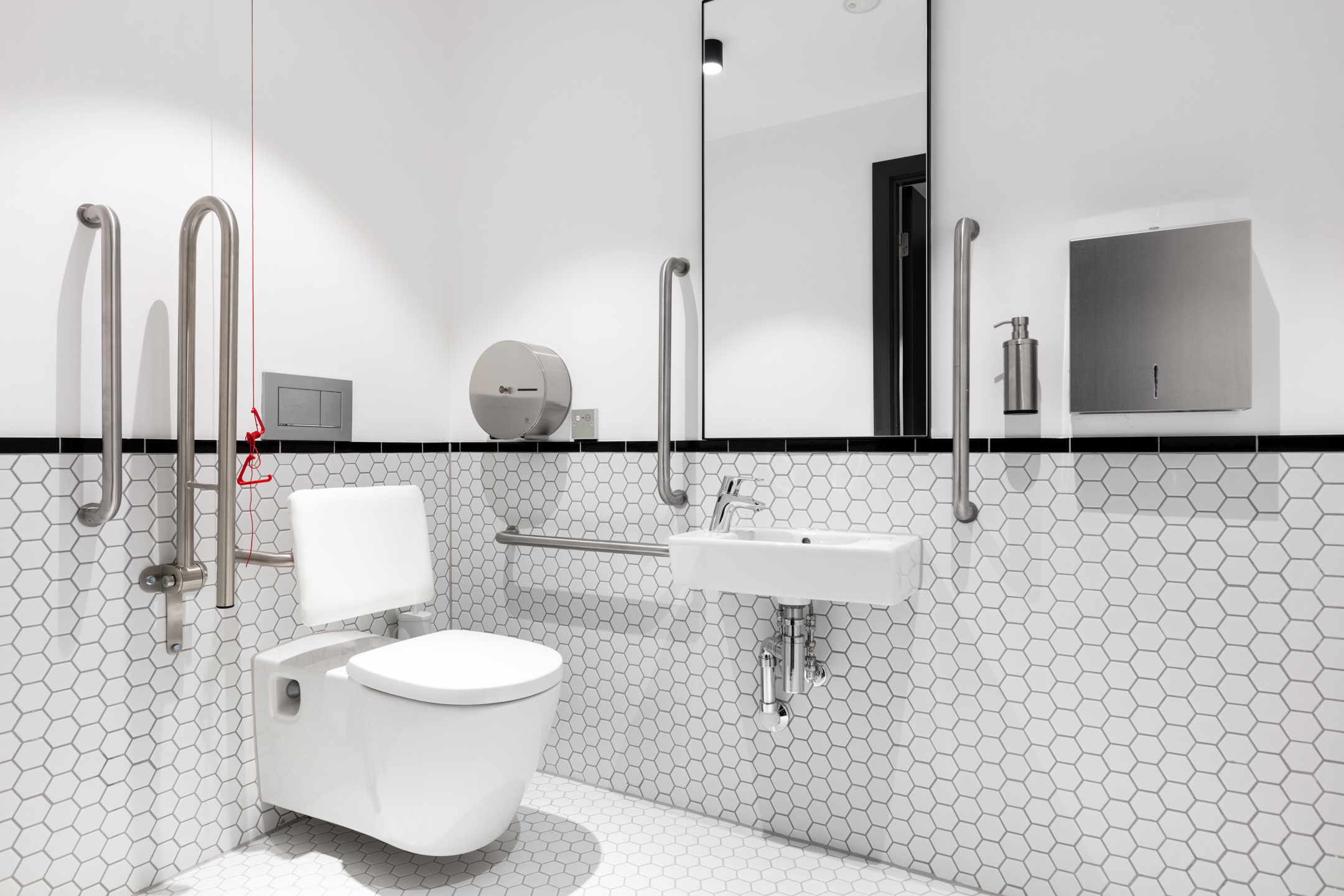 An accessible toilet in Programme Building with a minimalist design. The room features white hexagon tile walls, stainless steel grab rails, and a wall-mounted sink.