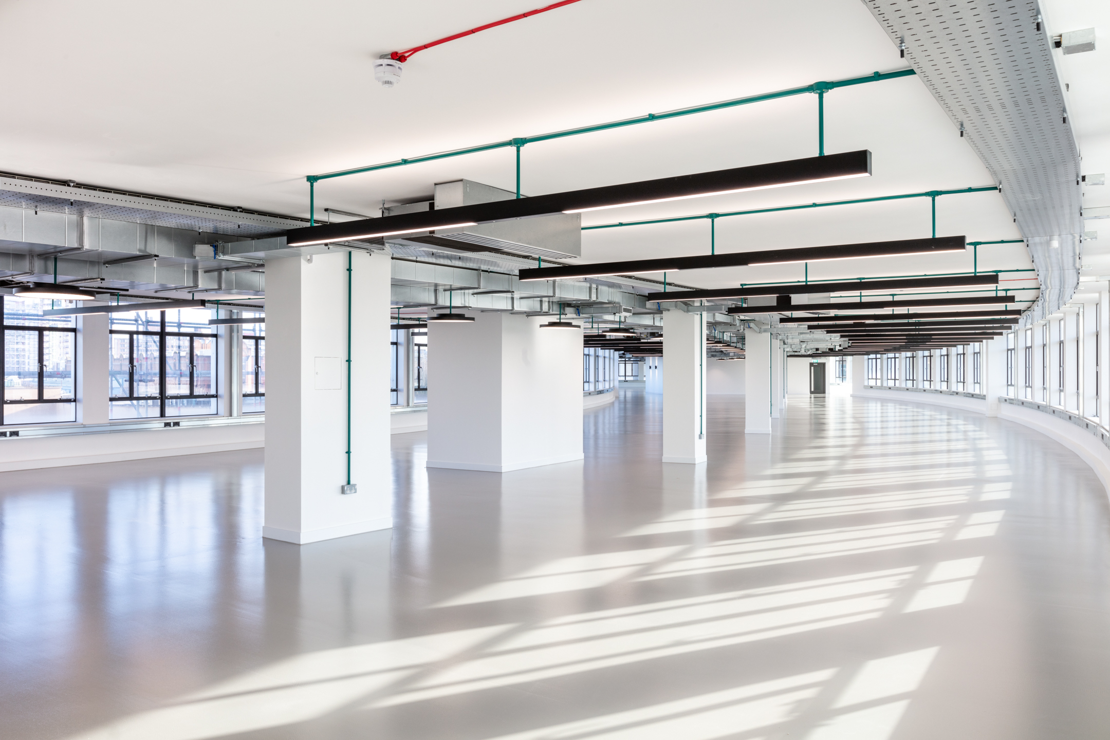 A bright, open floor at Programme Building, showcasing curved walls lined with floor-to-ceiling windows. Exposed ceiling structures and light fixtures contribute to the modern aesthetic.