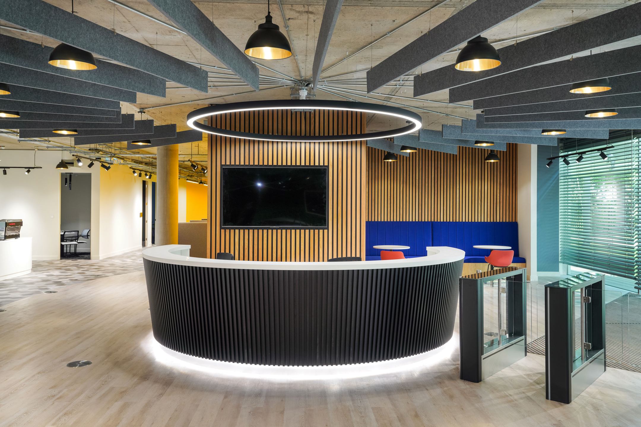 Reception area at Richmond University with a modern circular desk, wood panelling, acoustic ceiling baffles, and vibrant seating. A glass entry gate provides access, complemented by stylish pendant lighting.