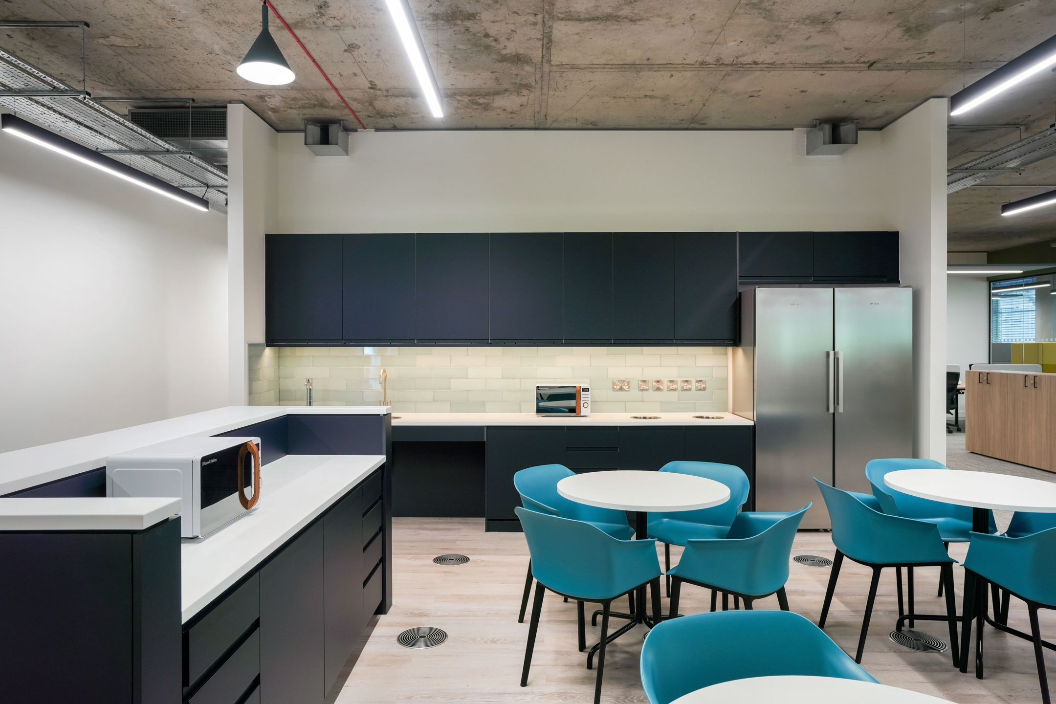 Teapoint area at Richmond University with sleek dark cabinetry, a tiled backsplash, stainless steel appliances, and round tables with teal chairs for seating.