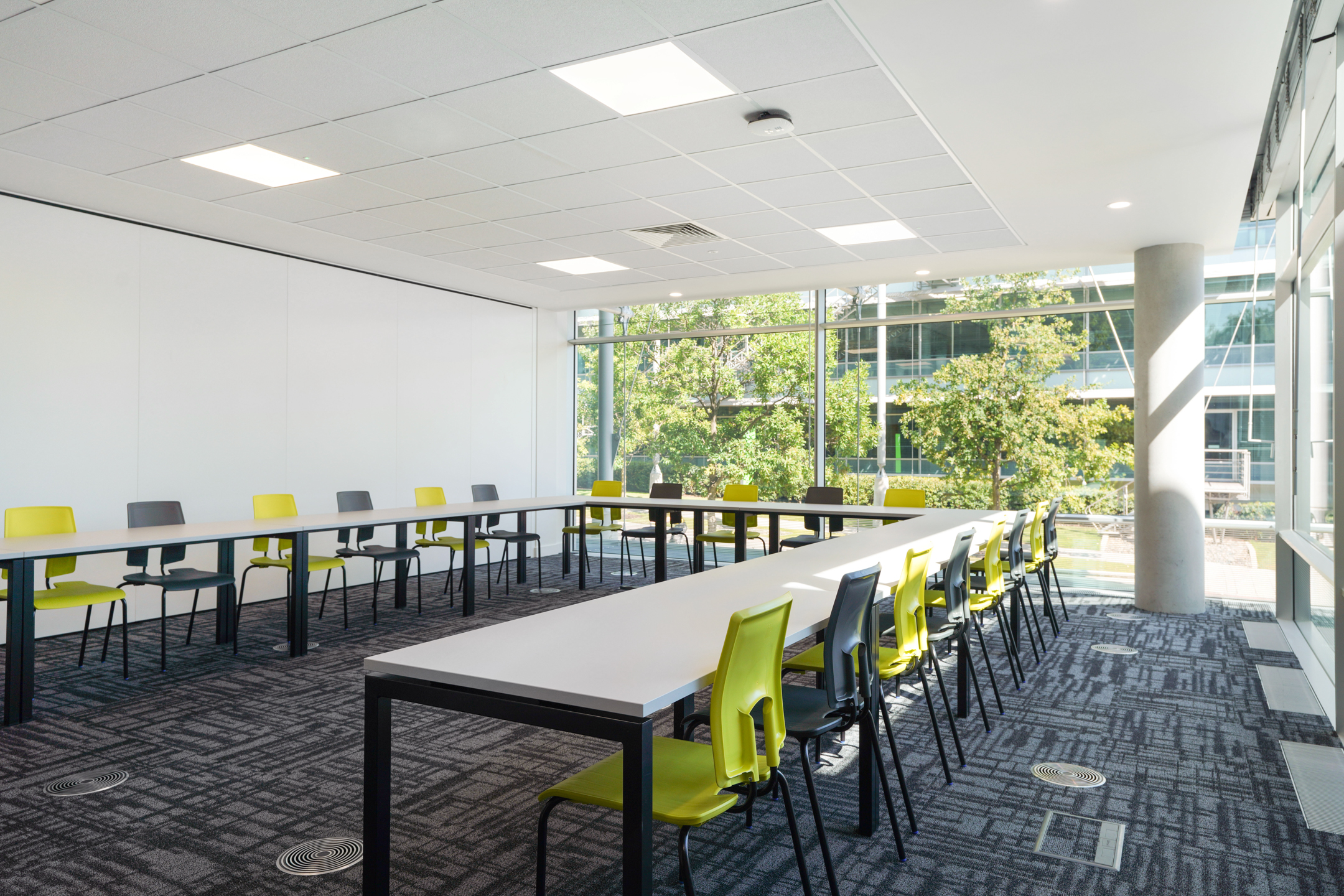 Bright classroom at Richmond University with large windows overlooking greenery, U-shaped table setup, and a mix of yellow and grey chairs arranged for collaborative learning.