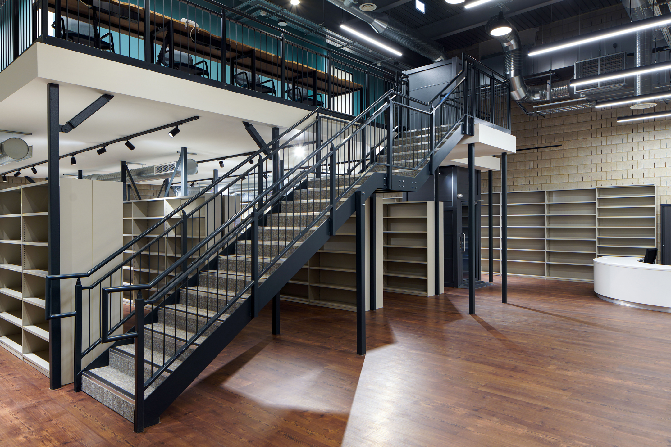 Library space at Richmond University with industrial-style metal staircase, mezzanine level seating, open shelving units, and exposed ductwork, complemented by wood flooring and ambient lighting.