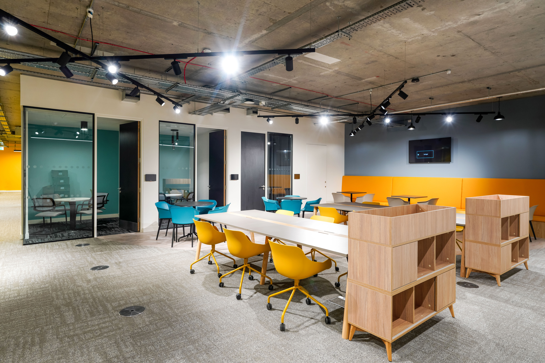 Modern study and collaboration area at Richmond University, featuring vibrant yellow and teal chairs, open shelving units, private meeting rooms with glass doors, and an exposed industrial ceiling with track lighting