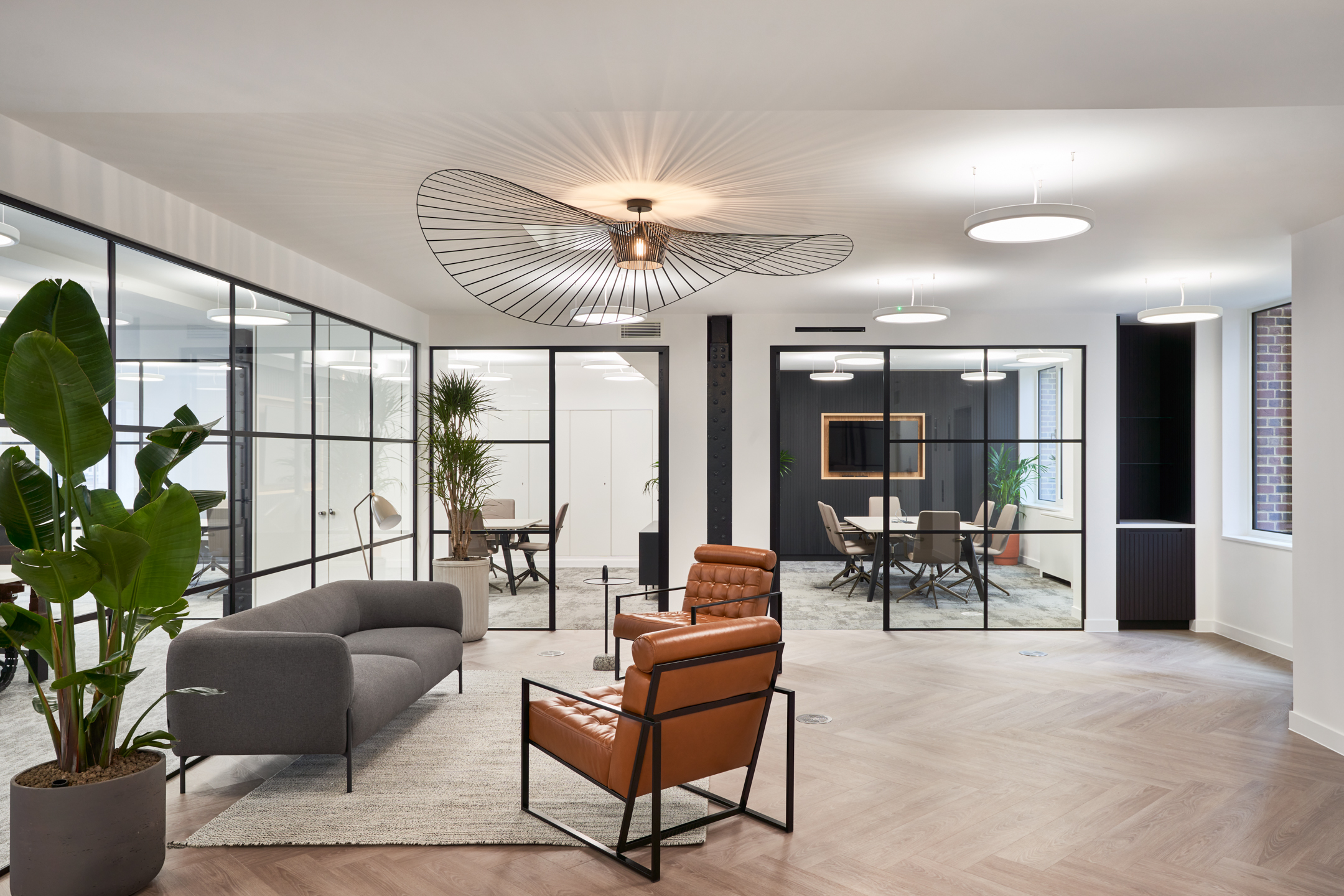 Breakout area at 3 St Helens Place with leather armchairs, a minimalist grey sofa, indoor plants, and glass-partitioned meeting rooms, creating a stylish and inviting workspace.