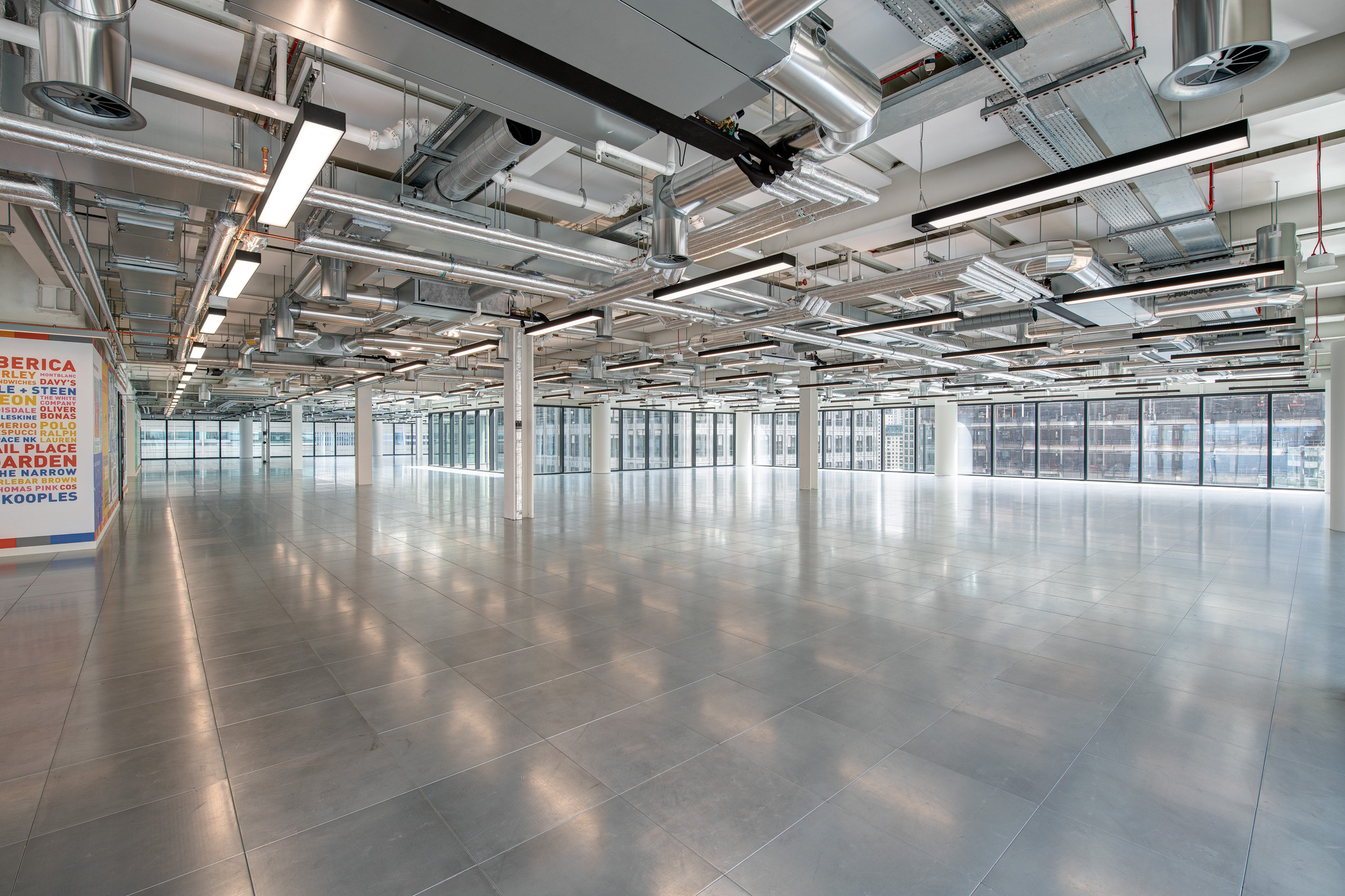 Open and expansive office space at 25 North Colonnade, showcasing reflective flooring, exposed industrial ceiling with ductwork, and large windows providing ample natural light.