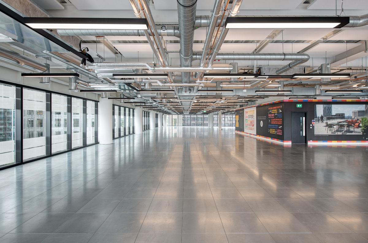 Spacious interior of 25 North Colonnade, showcasing exposed ductwork on the ceiling, floor-to-ceiling windows for natural light, and modern wall displays, creating an industrial yet polished office atmosphere.