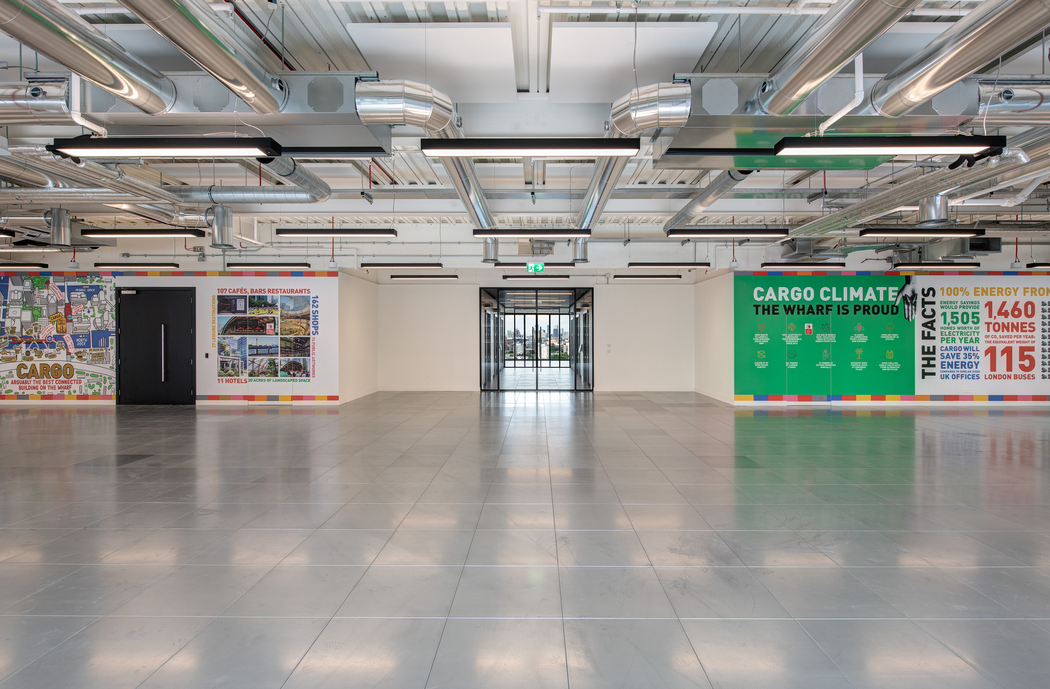 Interior view at 25 North Colonnade with colourful wall murals featuring graphics and environmental facts, exposed ceiling ducts, and reflective flooring, leading to a glass entrance.