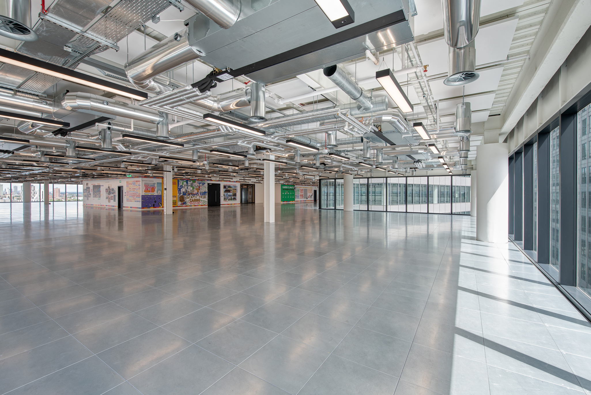 Bright open-plan workspace at 25 North Colonnade, highlighting exposed industrial ductwork, reflective flooring, large windows along one side, and vibrant wall displays.