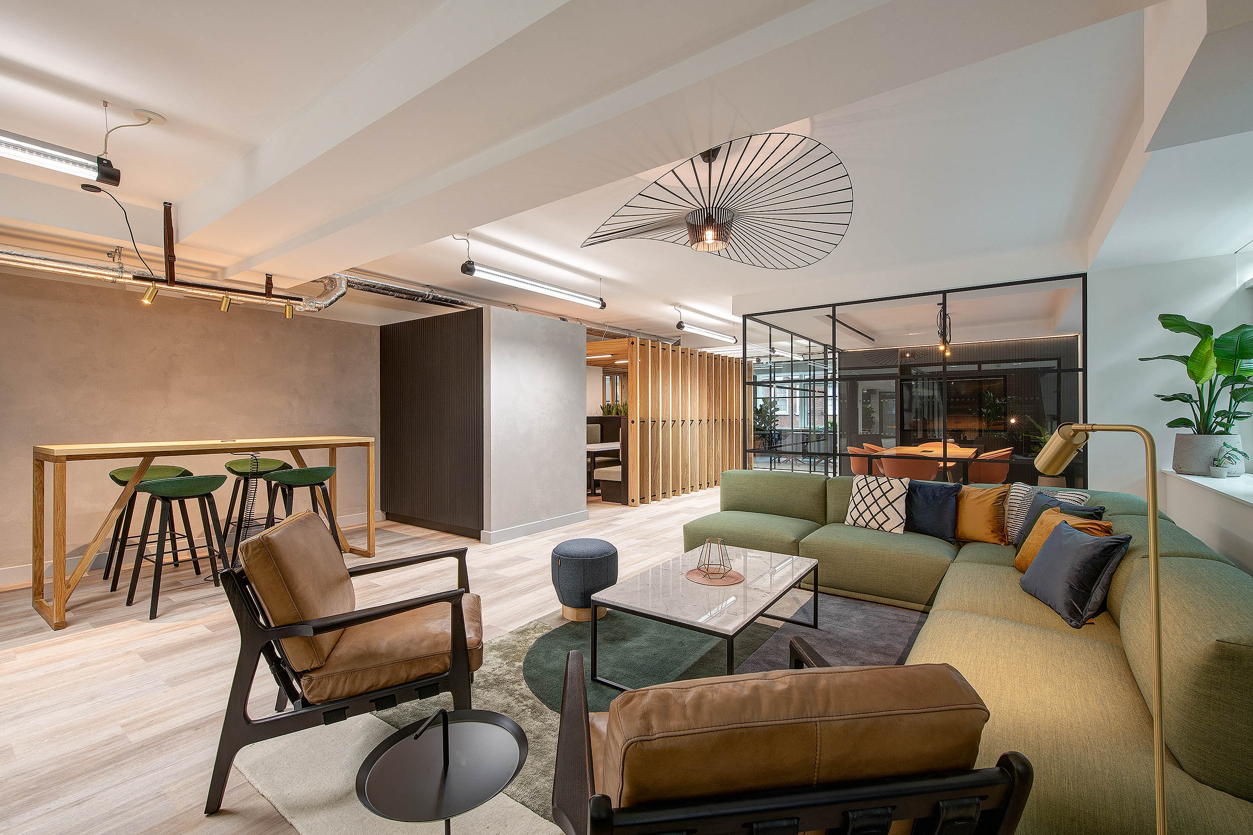 A cosy lounge area with a green L-shaped sofa, modern armchairs, and a marble coffee table, at 101 Cavendish Street. Industrial-style lighting and a wood panel feature.