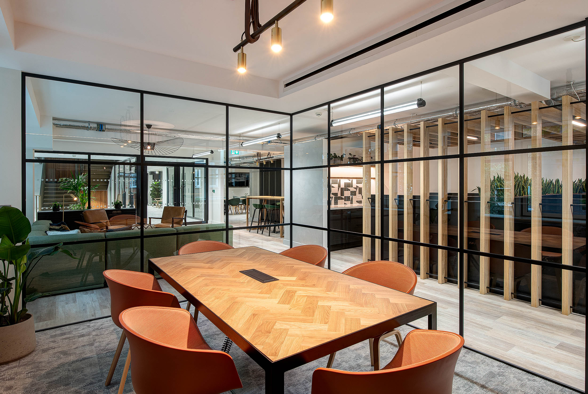 A modern meeting room with a wood herringbone table and orange chairs, surrounded by glass partitions and warm lighting, providing a view of an open lounge area.