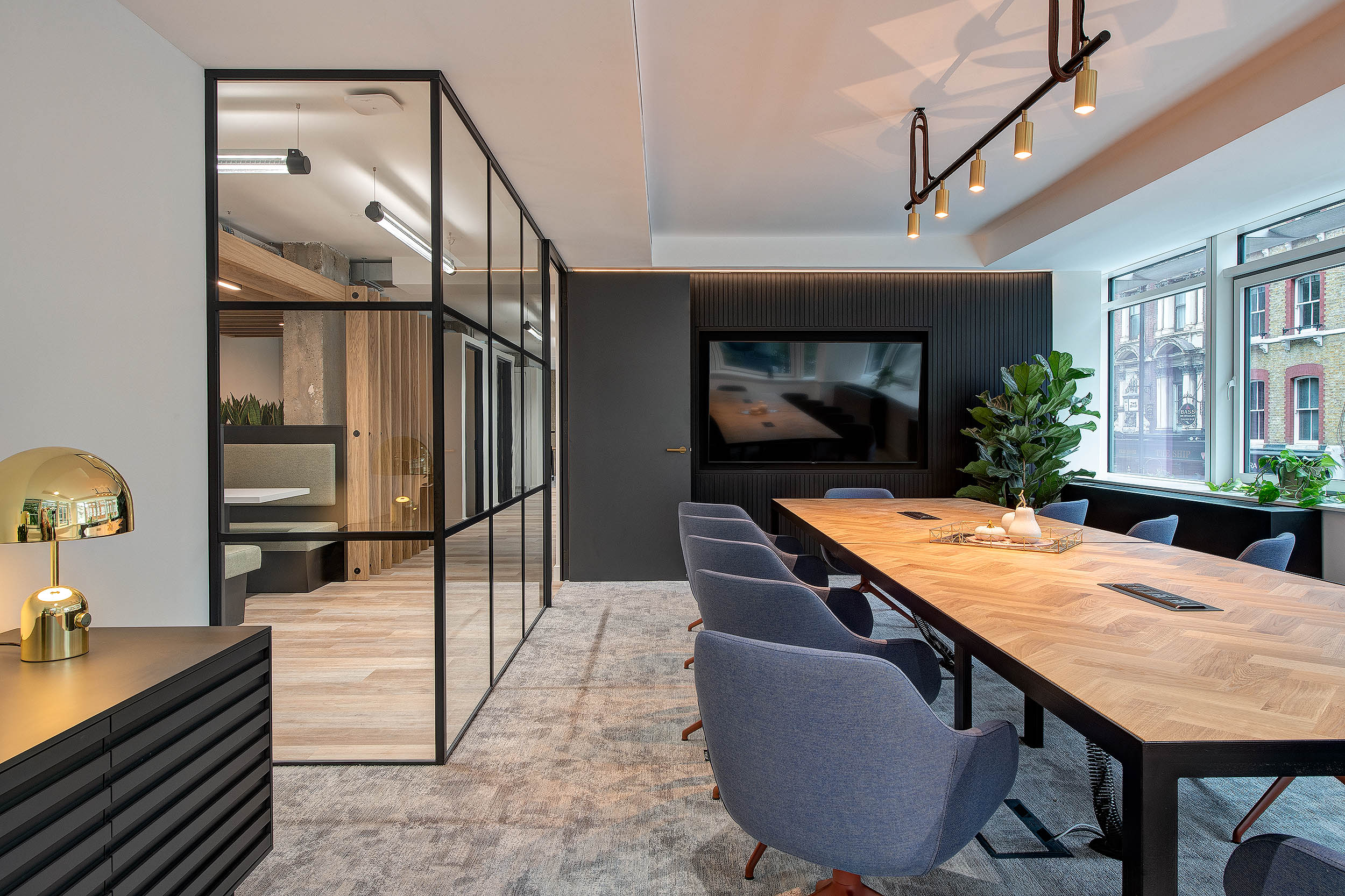 A stylish conference room at 101 New Cavendish Street, featuring a large wooden table with blue chairs, modern lighting fixtures, and glass walls, with a view into a private booth.