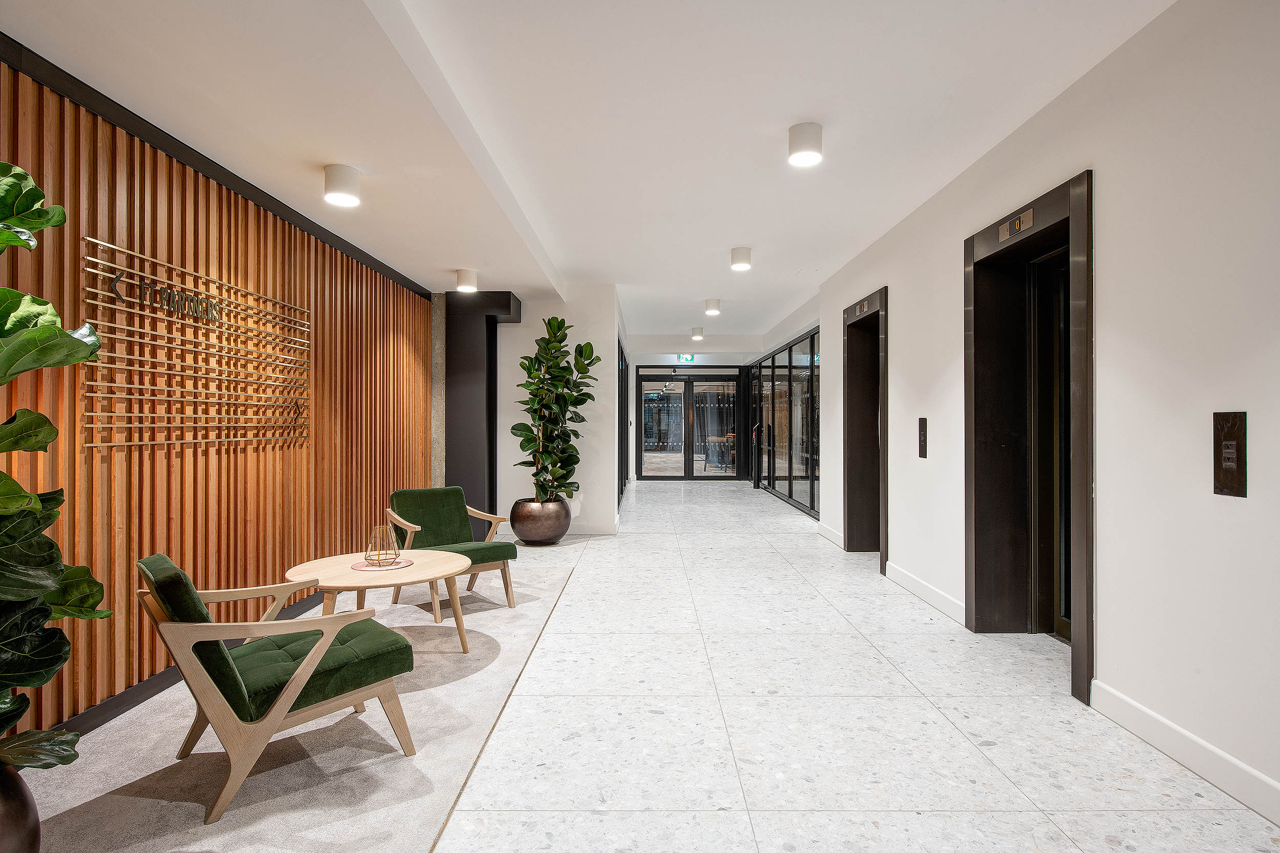 An open and welcoming office hallway at 101 New Cavendish Street, with a wood panel accent wall, green chairs, and plants. The clean and modern design is complemented by bright lighting.