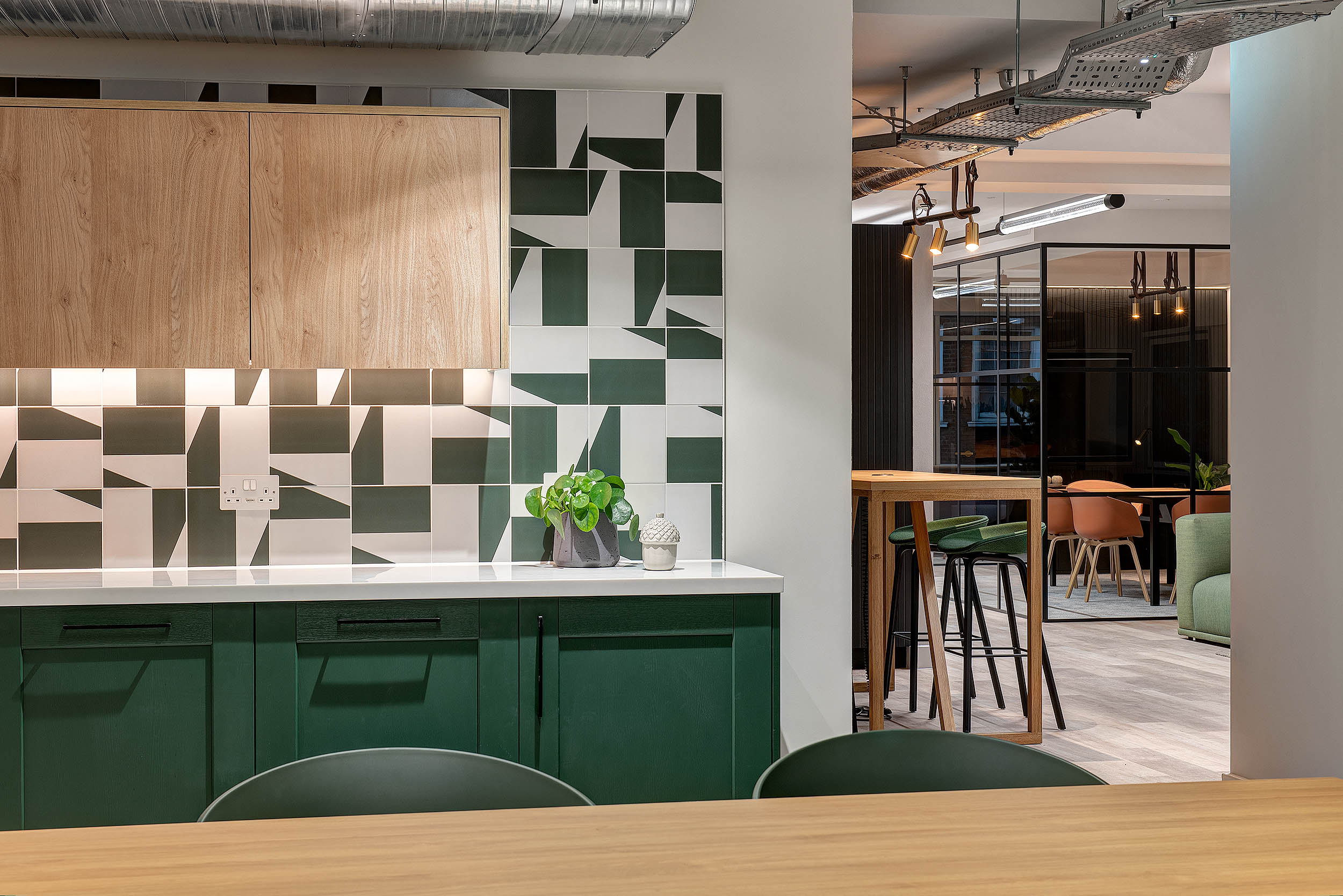 A teapoint area with a white countertop, green cabinetry, and geometric wall tiles in neutral colours. It leads to an open-plan lounge with a wood bar table and stools.