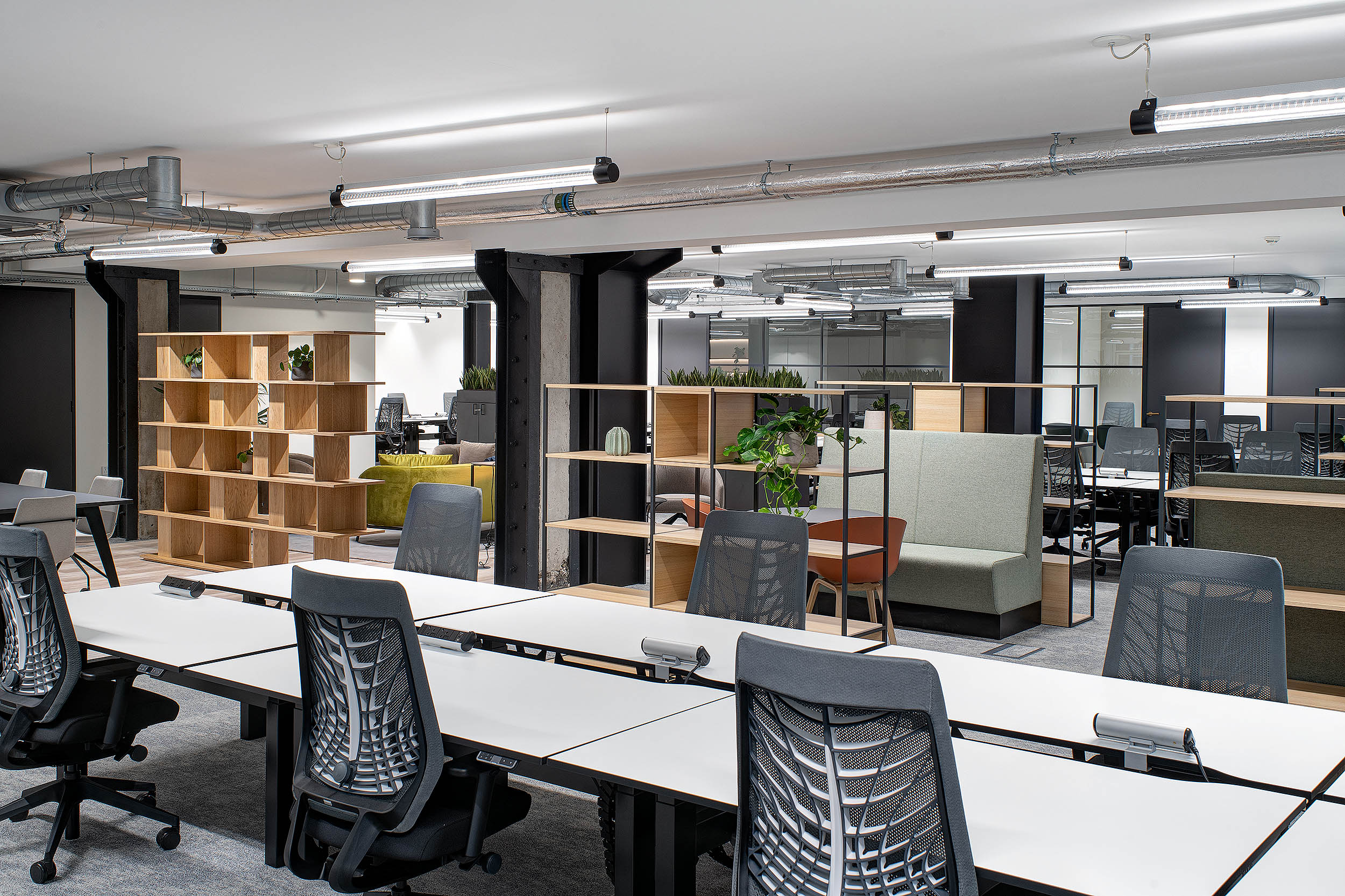 A spacious office floor featuring multiple white desks with ergonomic chairs, partitioned by wooden shelving and green booth seating areas.