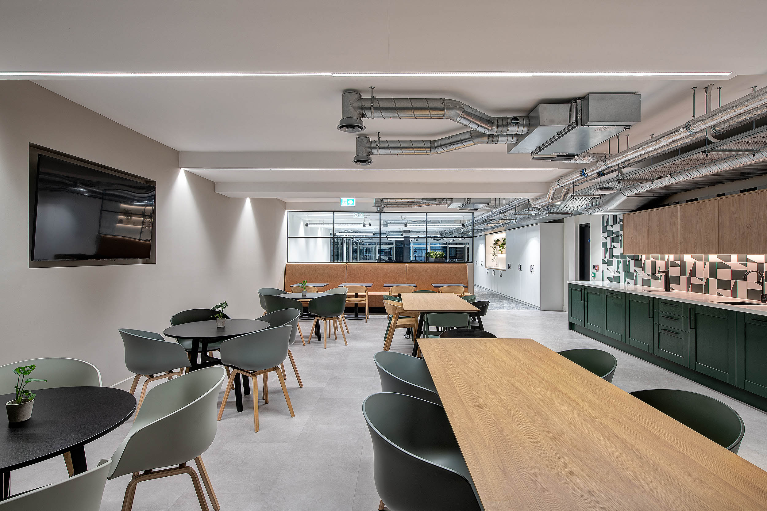 A large dining area with wooden tables and green chairs, complemented by sleek modern lighting. The space leads into a kitchen area with geometric tiles and cabinetry.