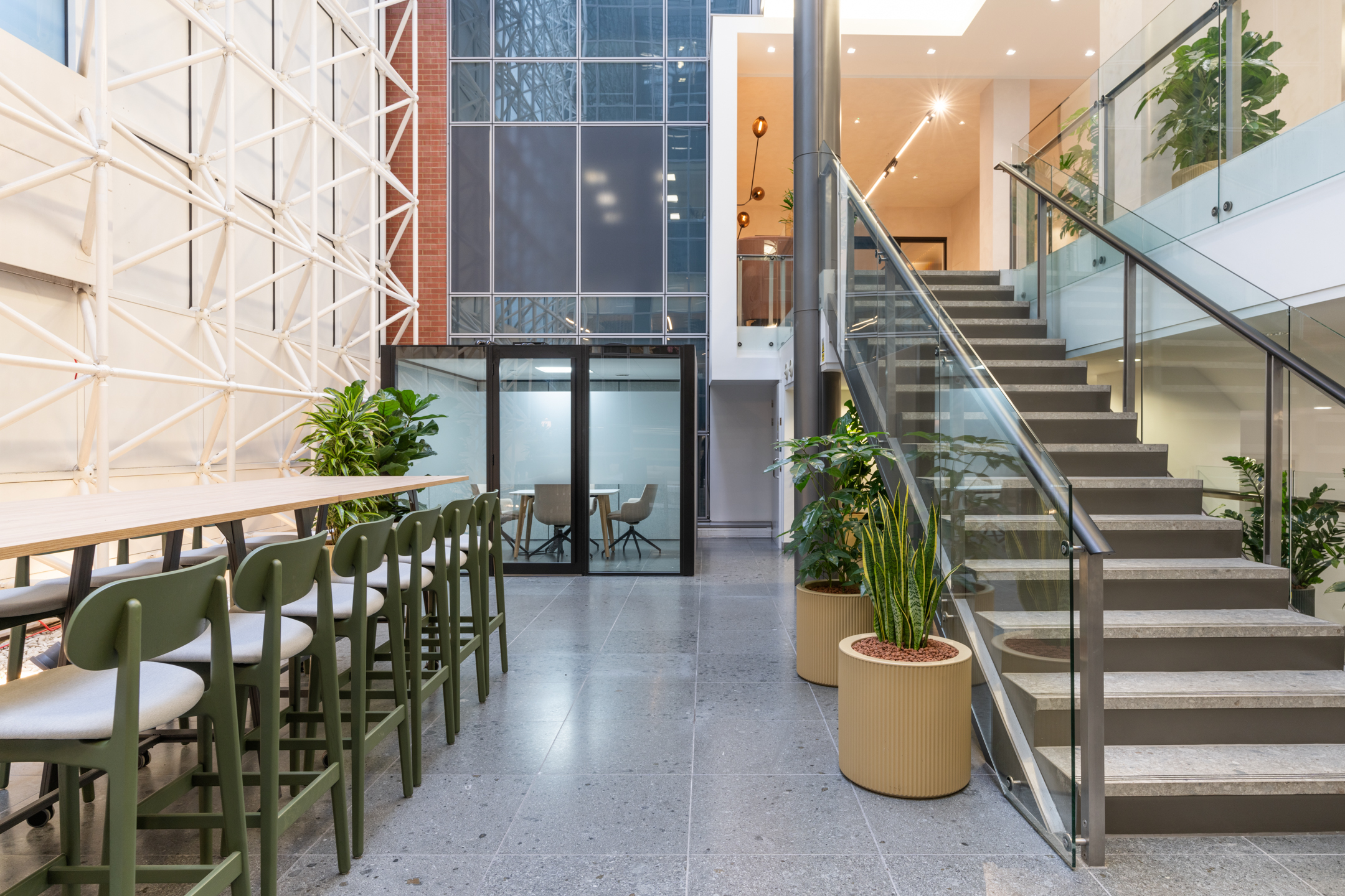 An open atrium space with a staircase on the right side and a seating area with green chairs and wooden furniture. The walls feature glass panels with an exterior metal framework visible outside.