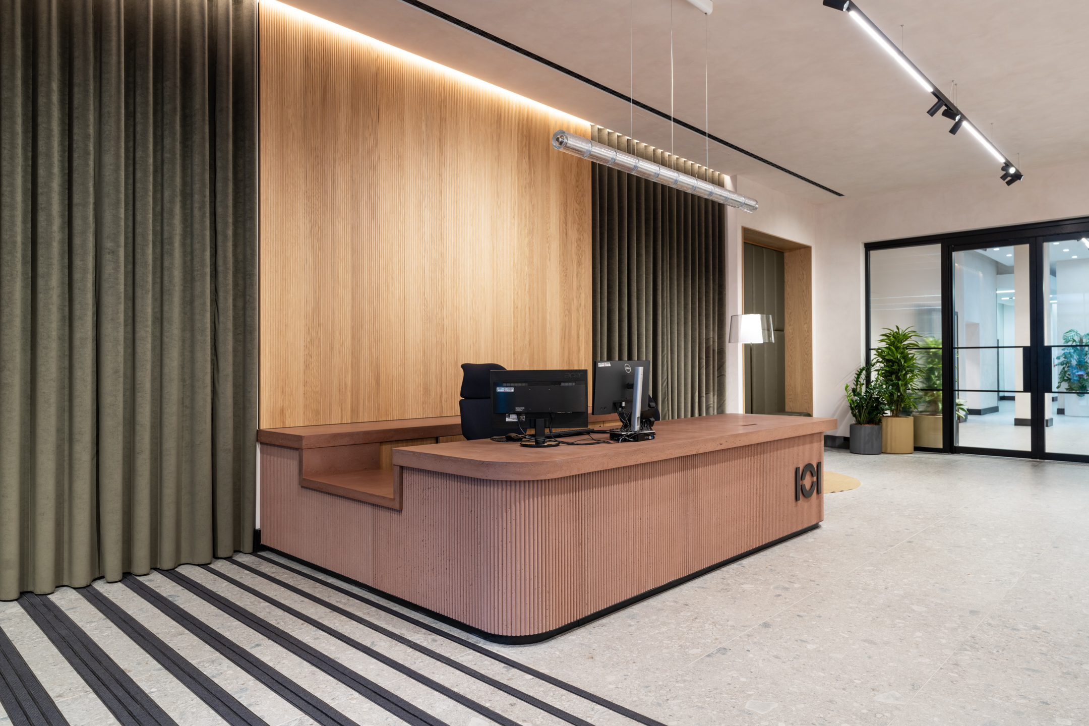 A stylish reception area with a wooden desk and dark accents, complemented by a curtain wall. The space is lit by hanging linear lights and features plants as decorative elements.