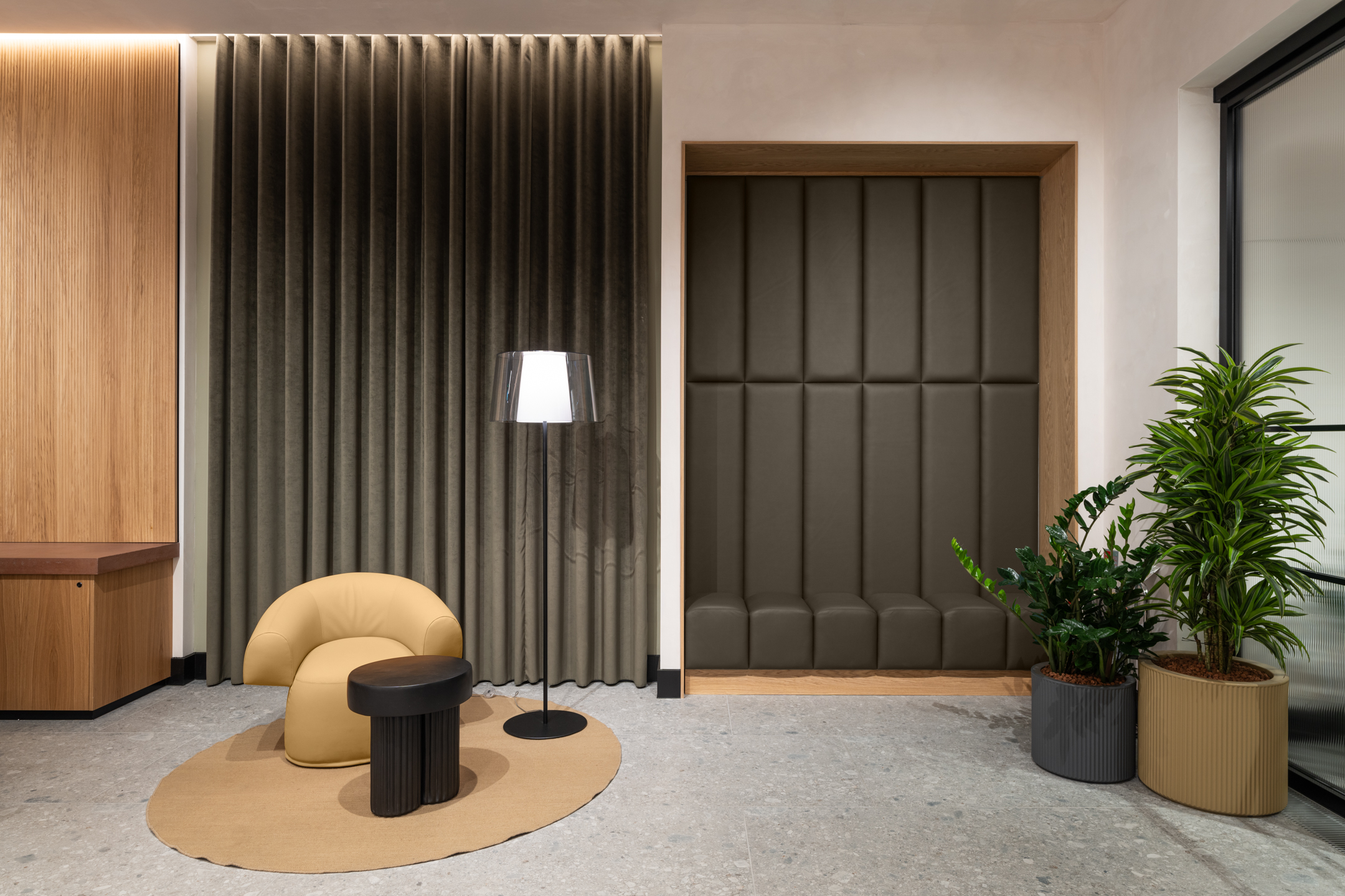 A cosy seating area with a beige armchair, small black side table, and modern standing lamp. The setting includes an olive-green padded wall and matching plants for a natural touch.