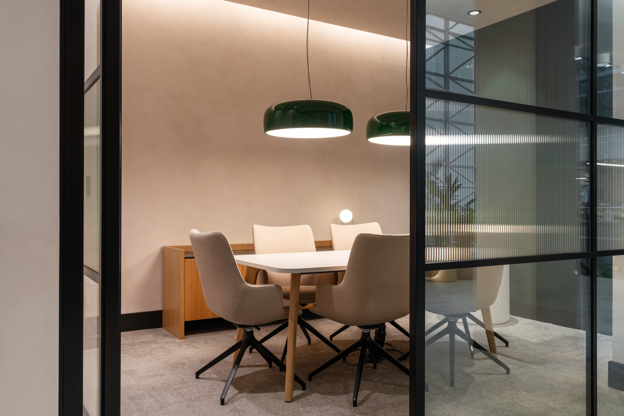 An enclosed meeting room at 101 St Martin's Lane with a round table surrounded by beige chairs and two pendant lights above. The room is framed with glass walls, allowing partial visibility from the corridor.