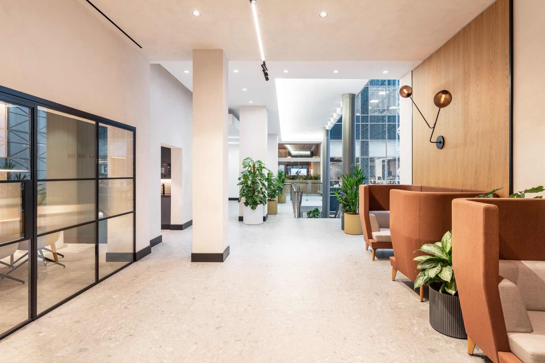 A hallway leading to an atrium, featuring booth seating with brown upholstery along one wall and decorative plants. The flooring is light-coloured, contrasting with the darker walls and furniture.