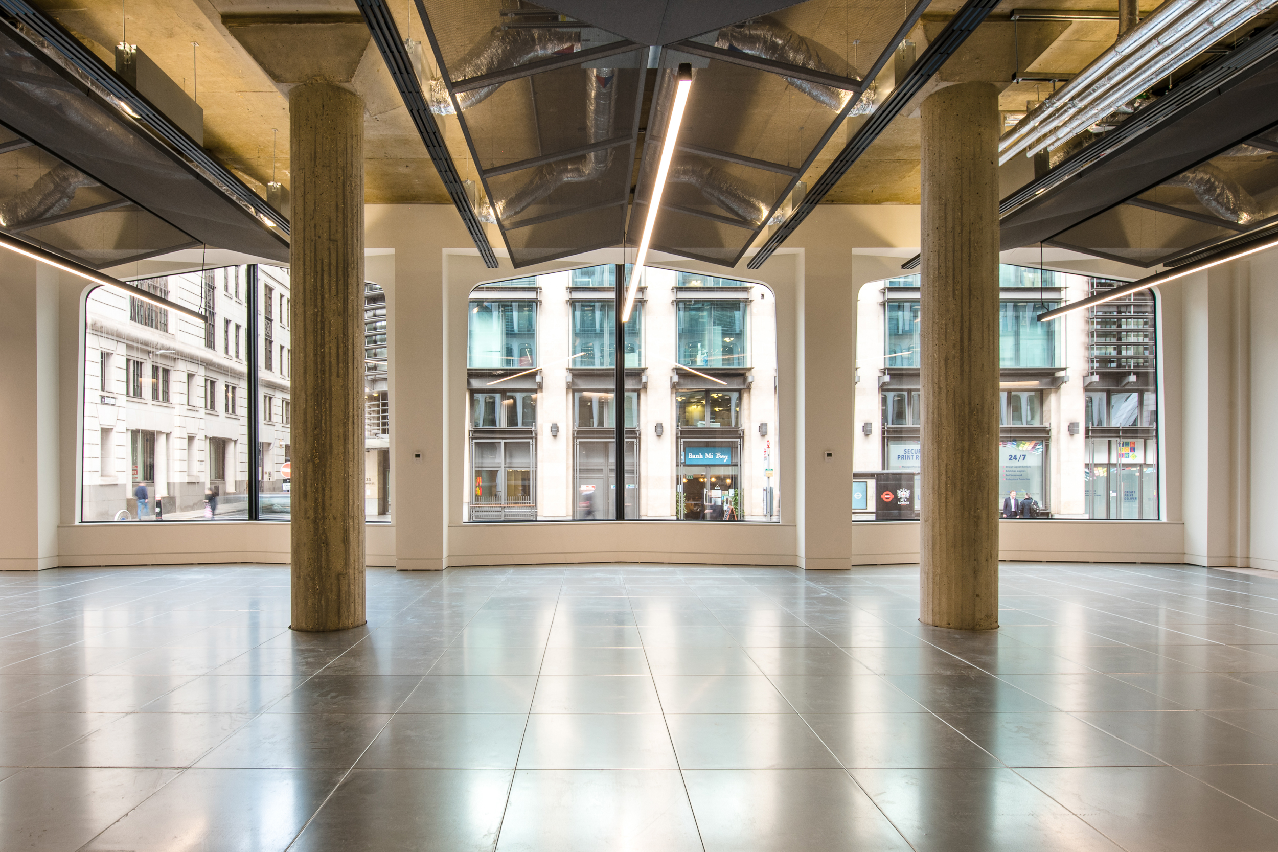 The image shows a spacious room with large windows, allowing natural light to flood the space. The interior features concrete pillars, exposed ceilings with ductwork, and reflective tiled flooring. Outside, a commercial building can be seen.