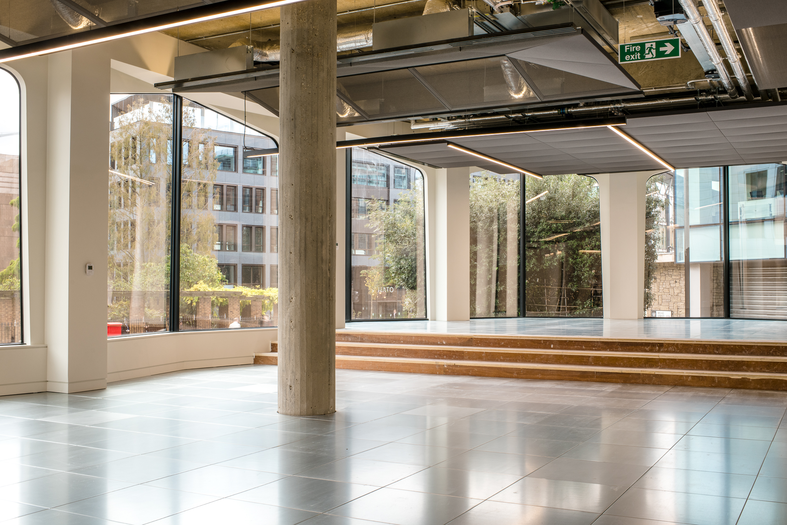 The image shows a bright interior with tiled floors, a small flight of steps leading to a raised section, and floor-to-ceiling windows offering an outdoor view. Exposed ductwork and ceiling lights complete the modern look.