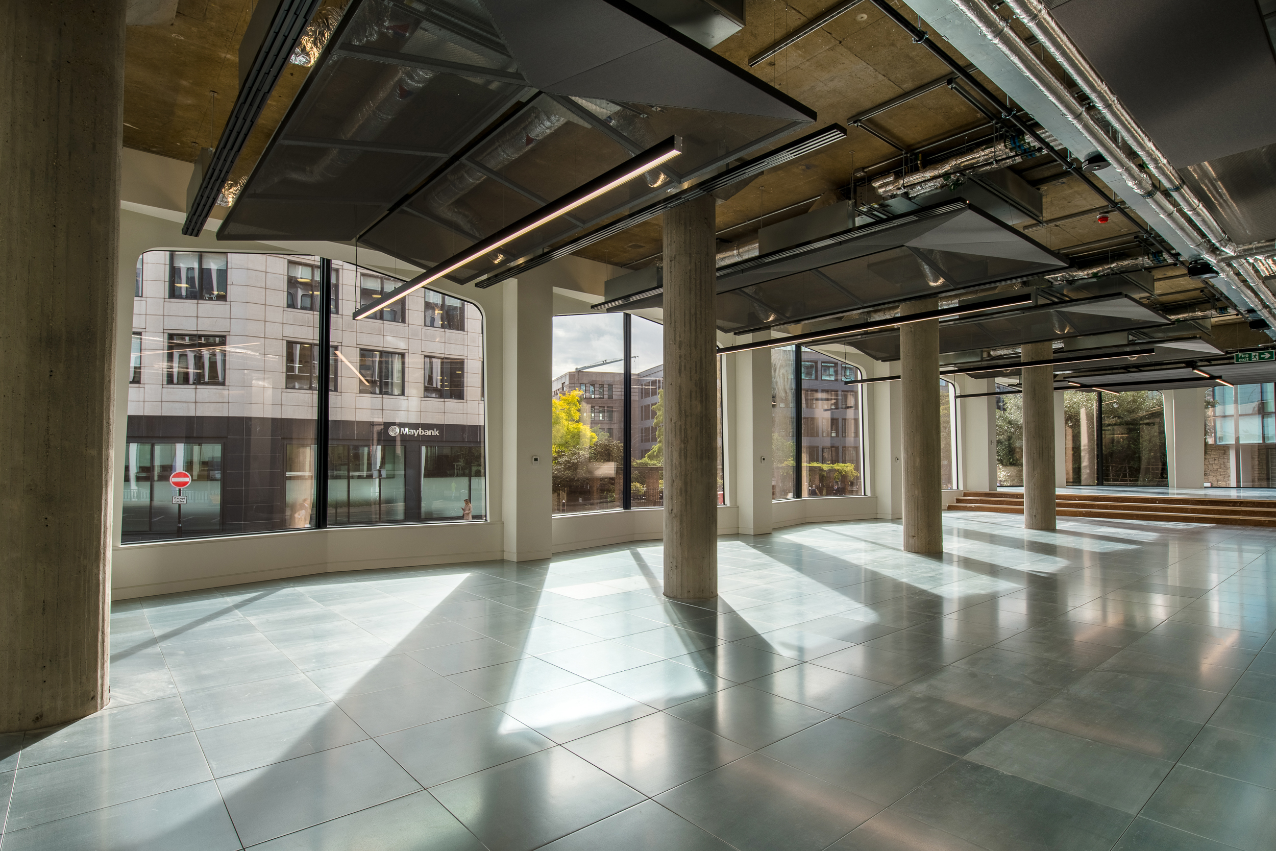 The image shows a spacious room with large windows, allowing natural light to flood the space. The interior features concrete pillars, exposed ceilings with ductwork, and reflective tiled flooring. Outside, a commercial building can be seen.