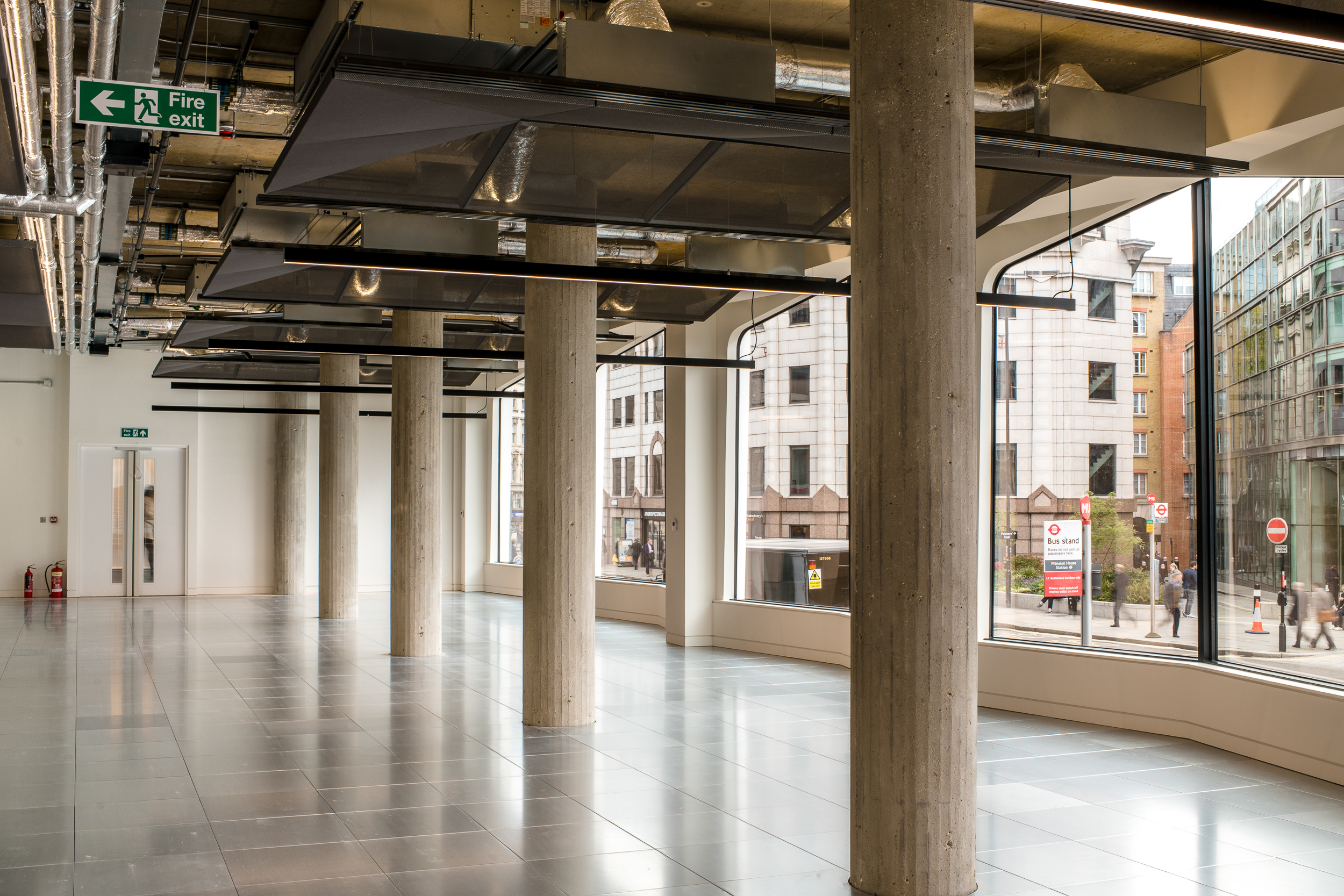 A bright, open space with polished tiled floors, concrete support columns, and large arched windows. The view outside shows urban surroundings with people walking and buildings in the background.