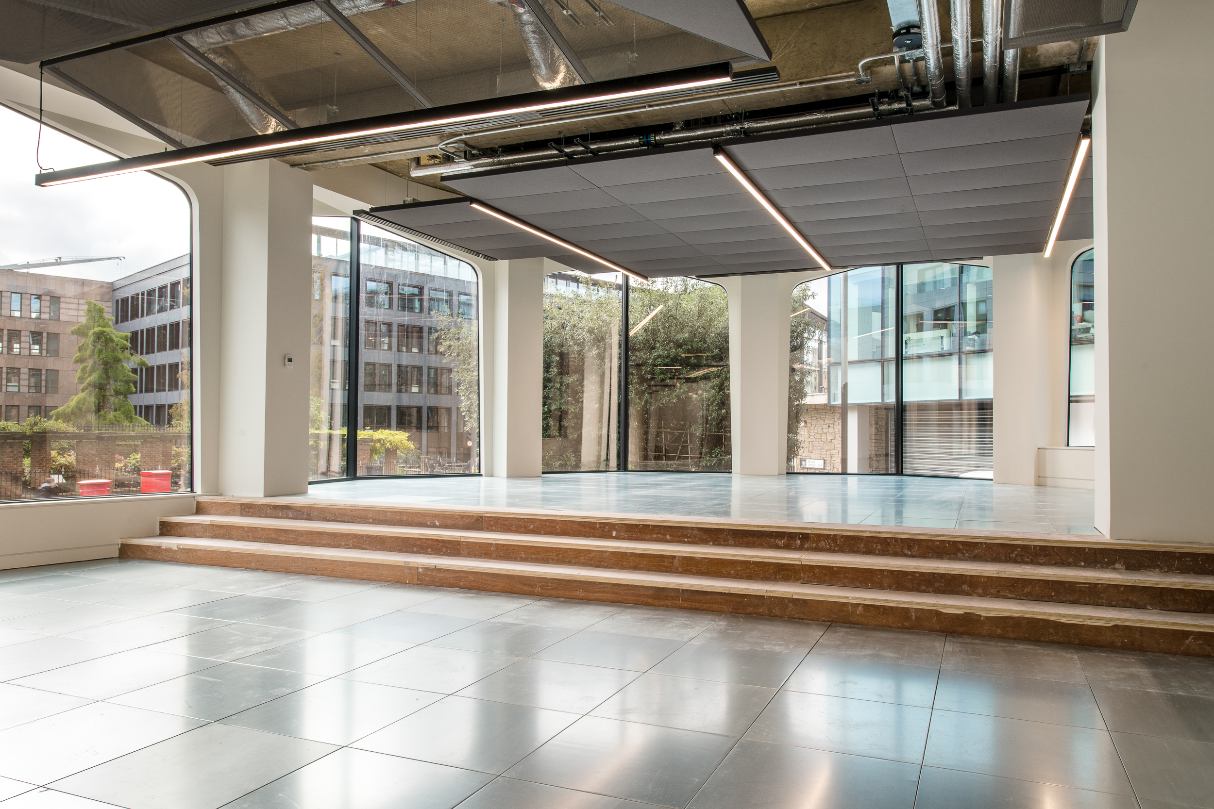 The image shows a bright interior with tiled floors, a small flight of steps leading to a raised section, and floor-to-ceiling windows offering an outdoor view. Exposed ductwork and ceiling lights complete the modern look.